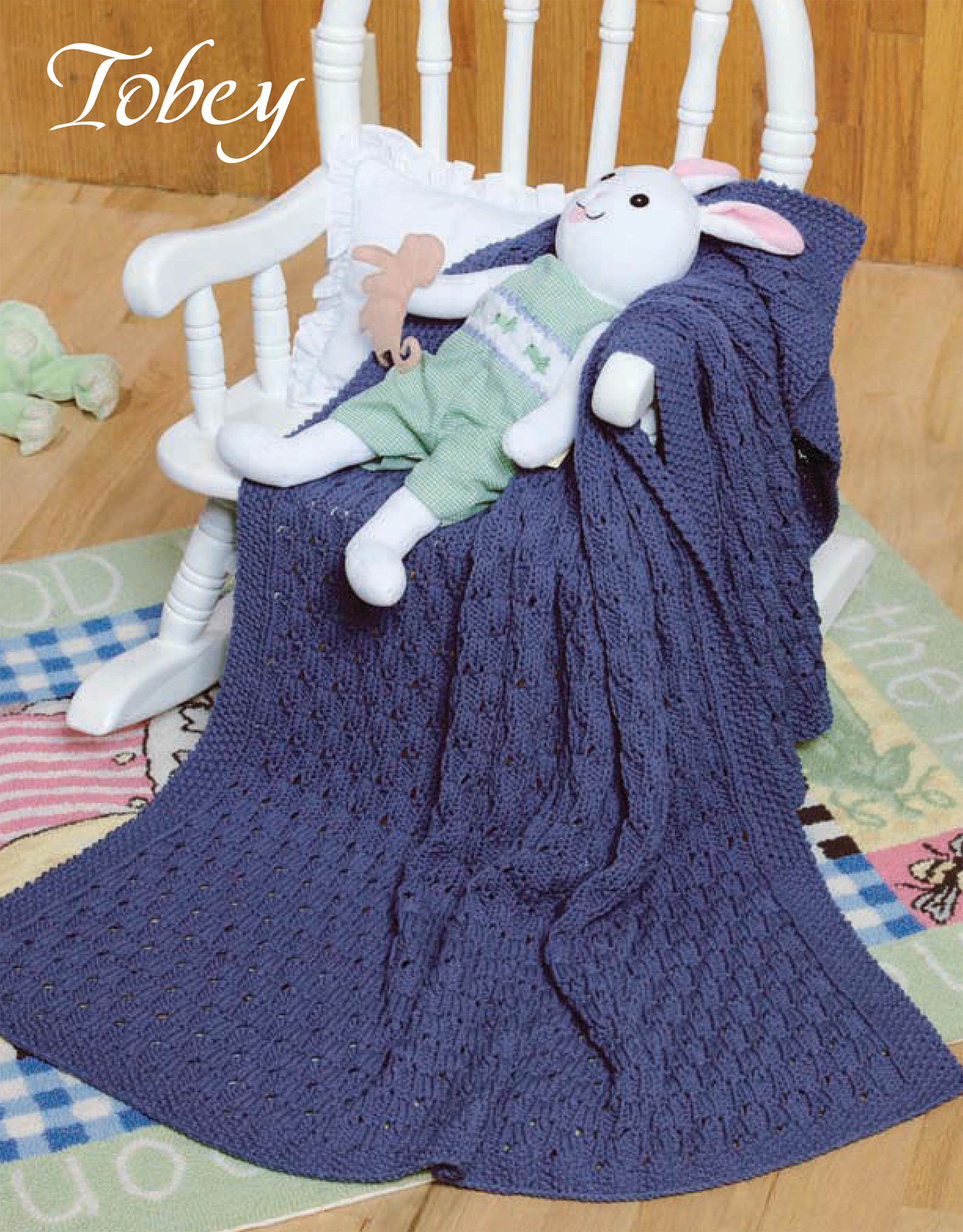 A white rocking chair with a plush rabbit in green rests by a blue blanket featuring a beginner pattern from "Quick Knit Keepsakes, Book 2" by Leisure Arts. The chair sits on a colorful rug, with "Tobey" in the top left corner.