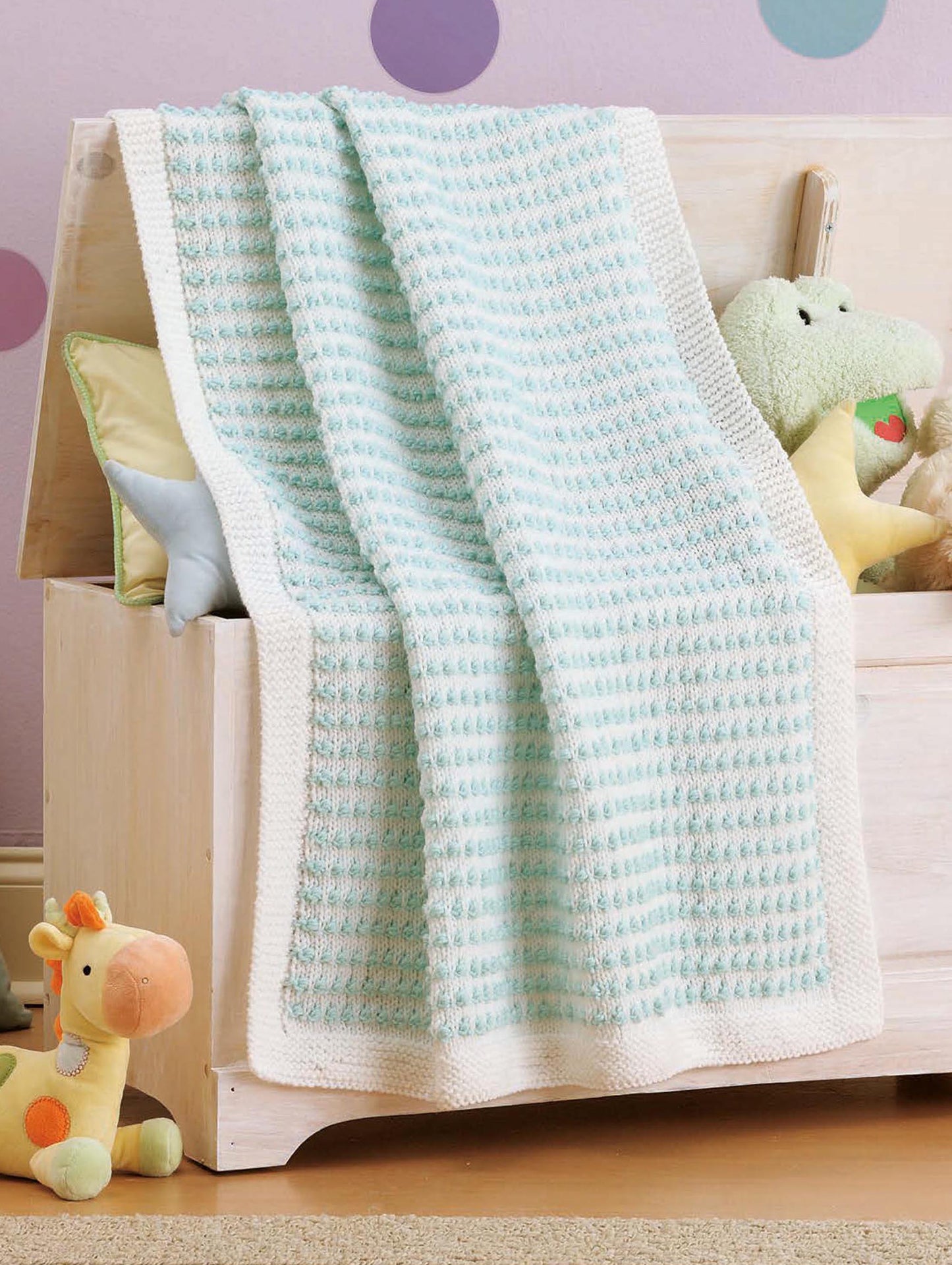 The Little Lullabies—Easy Blankets with Beautiful Color Patterns! by Leisure Arts is a soft, light blue and white knitted baby blanket, ideal for beginners, draped over a toy chest in a pastel nursery with plush toys.
