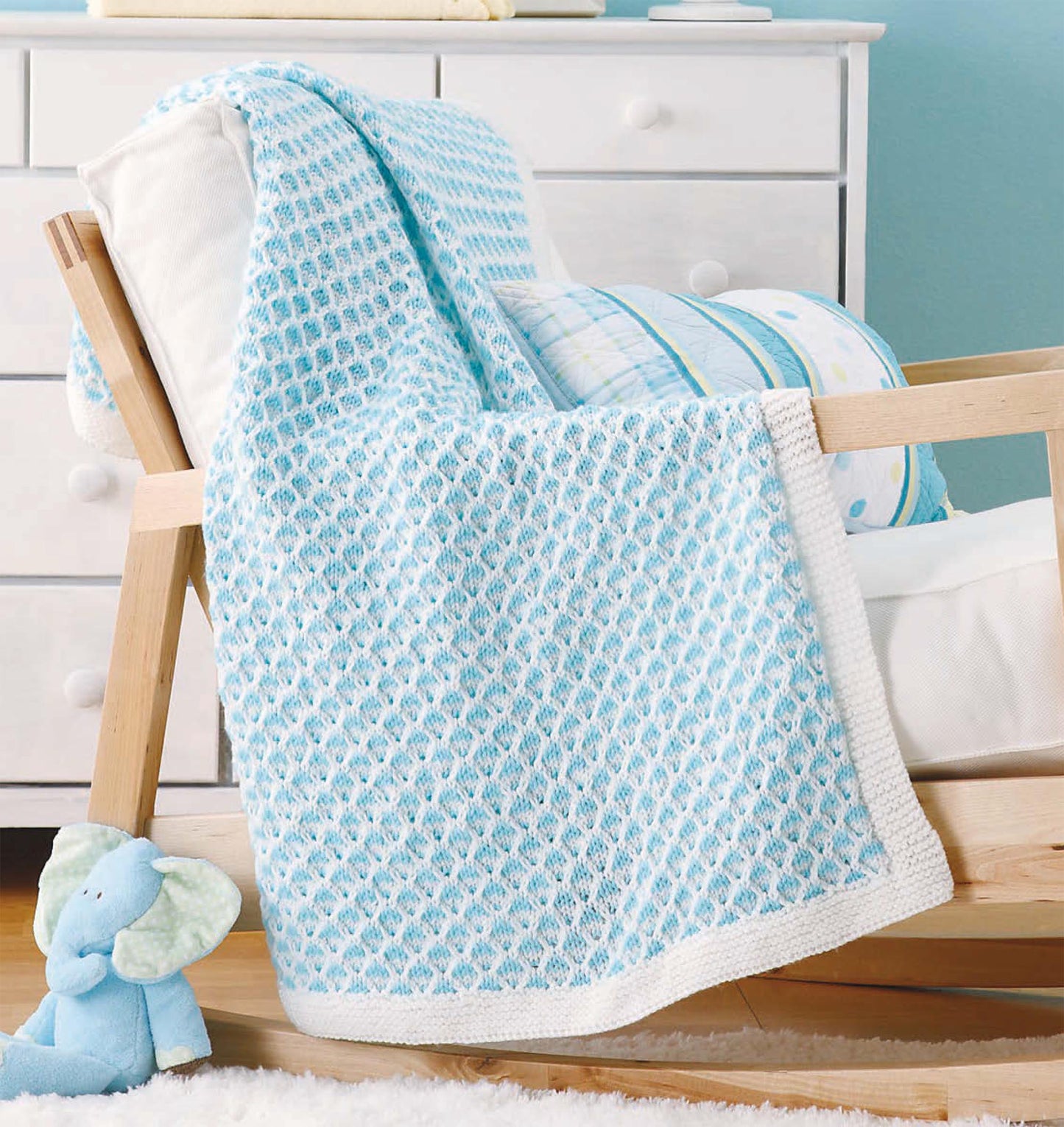 The Little Lullabies—Easy Blankets with Beautiful Color Patterns! by Leisure Arts, in light blue and white, is draped over a cushioned wooden rocking chair, with a stuffed blue elephant nearby for a cozy nursery look.