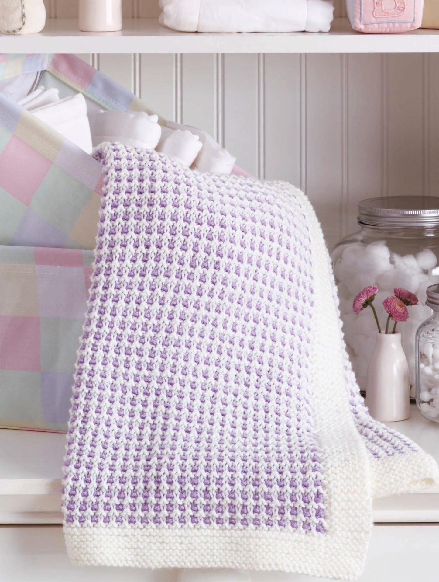 The Little Lullabies—Easy Blankets with Beautiful Color Patterns! by Leisure Arts, hand-knitted in purple and white with a white border, is draped over a pastel basket. Glass jars of cotton balls and pink flowers sit close by.