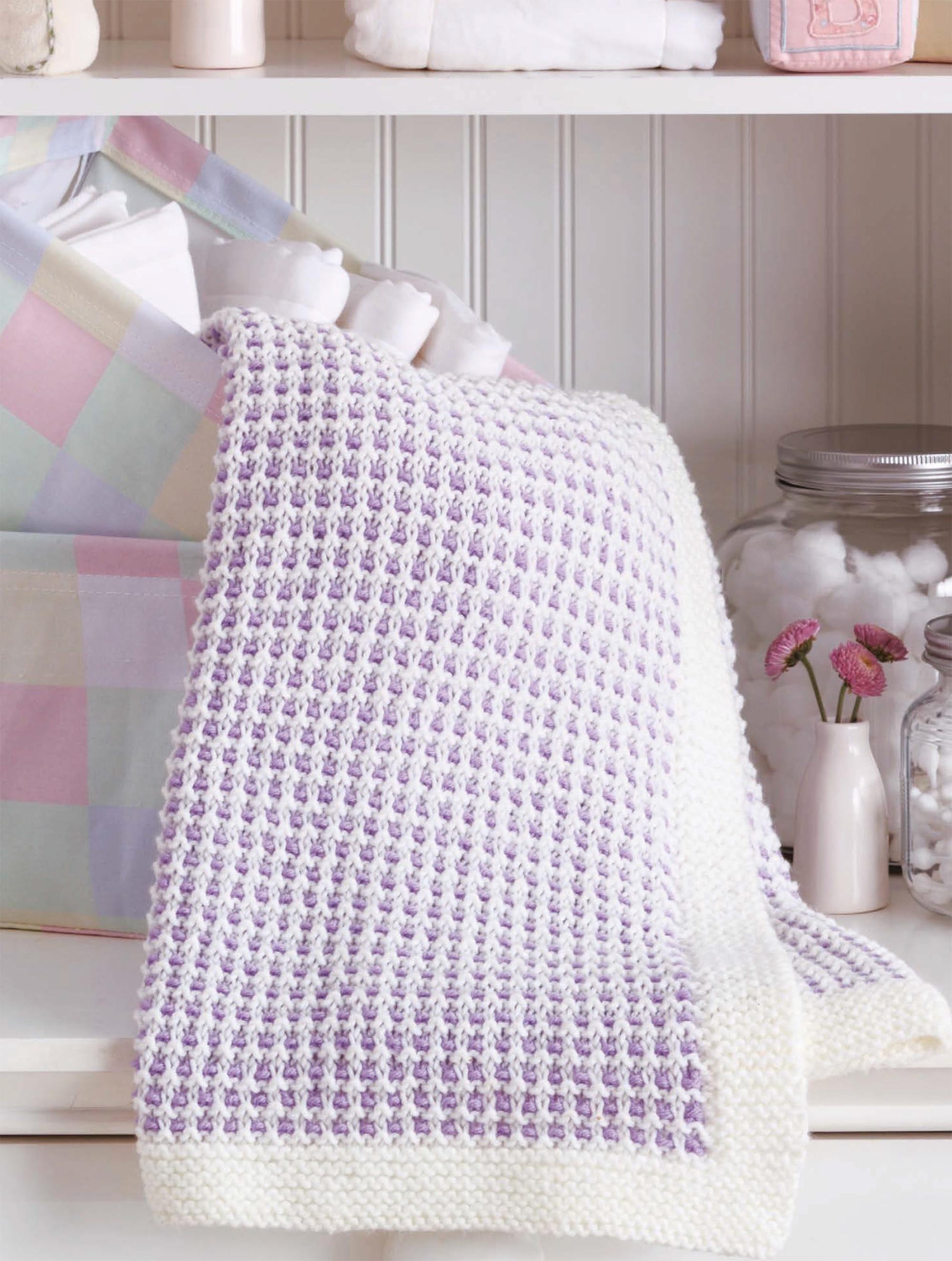 The Little Lullabies—Easy Blankets with Beautiful Color Patterns! by Leisure Arts, hand-knitted in purple and white with a white border, is draped over a pastel basket. Glass jars of cotton balls and pink flowers sit close by.
