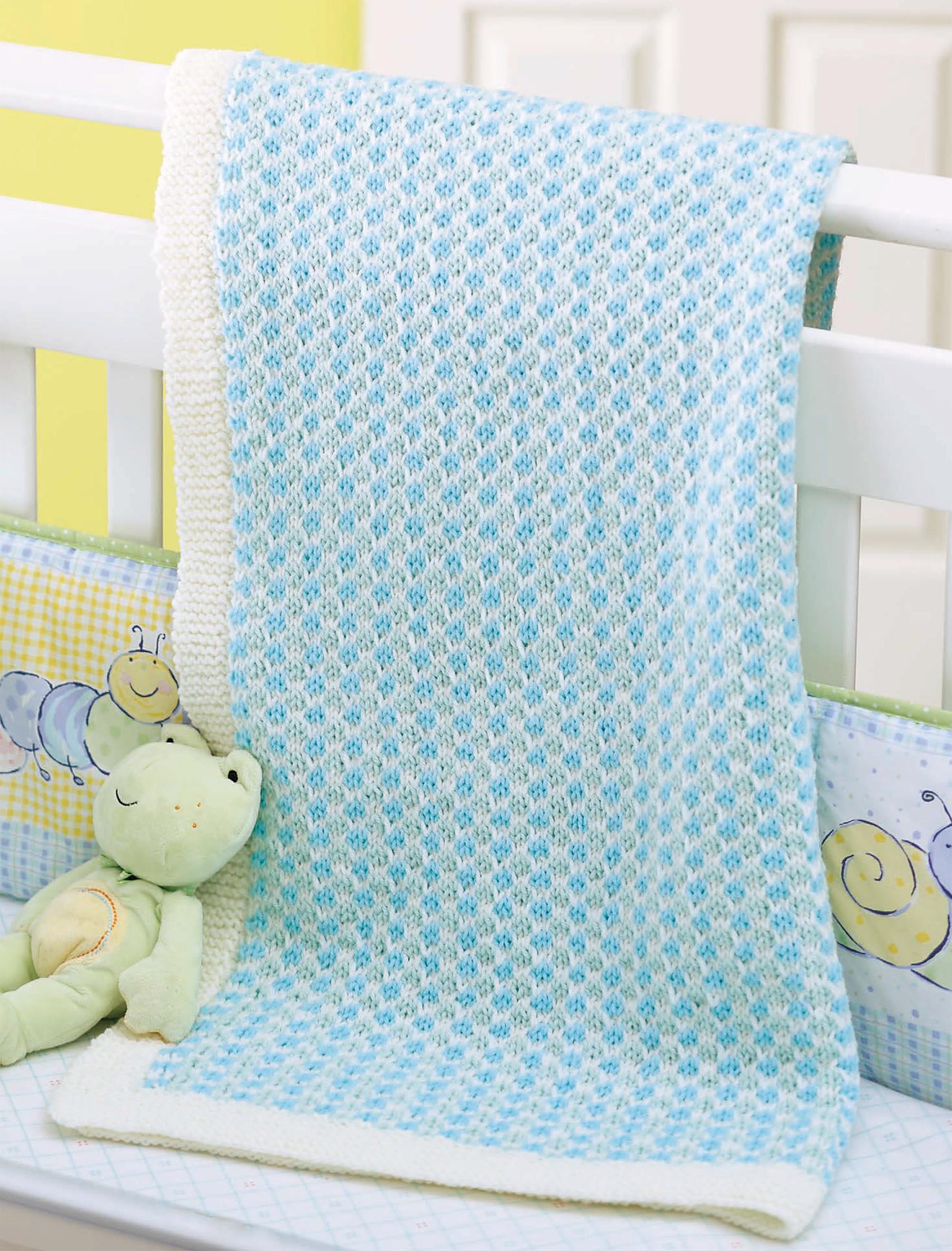 The Little Lullabies—Easy Blankets with Beautiful Color Patterns! by Leisure Arts, a soft light blue and white knit, hangs over the white crib next to a green plush frog and crib bumpers with smiling caterpillars and snails.