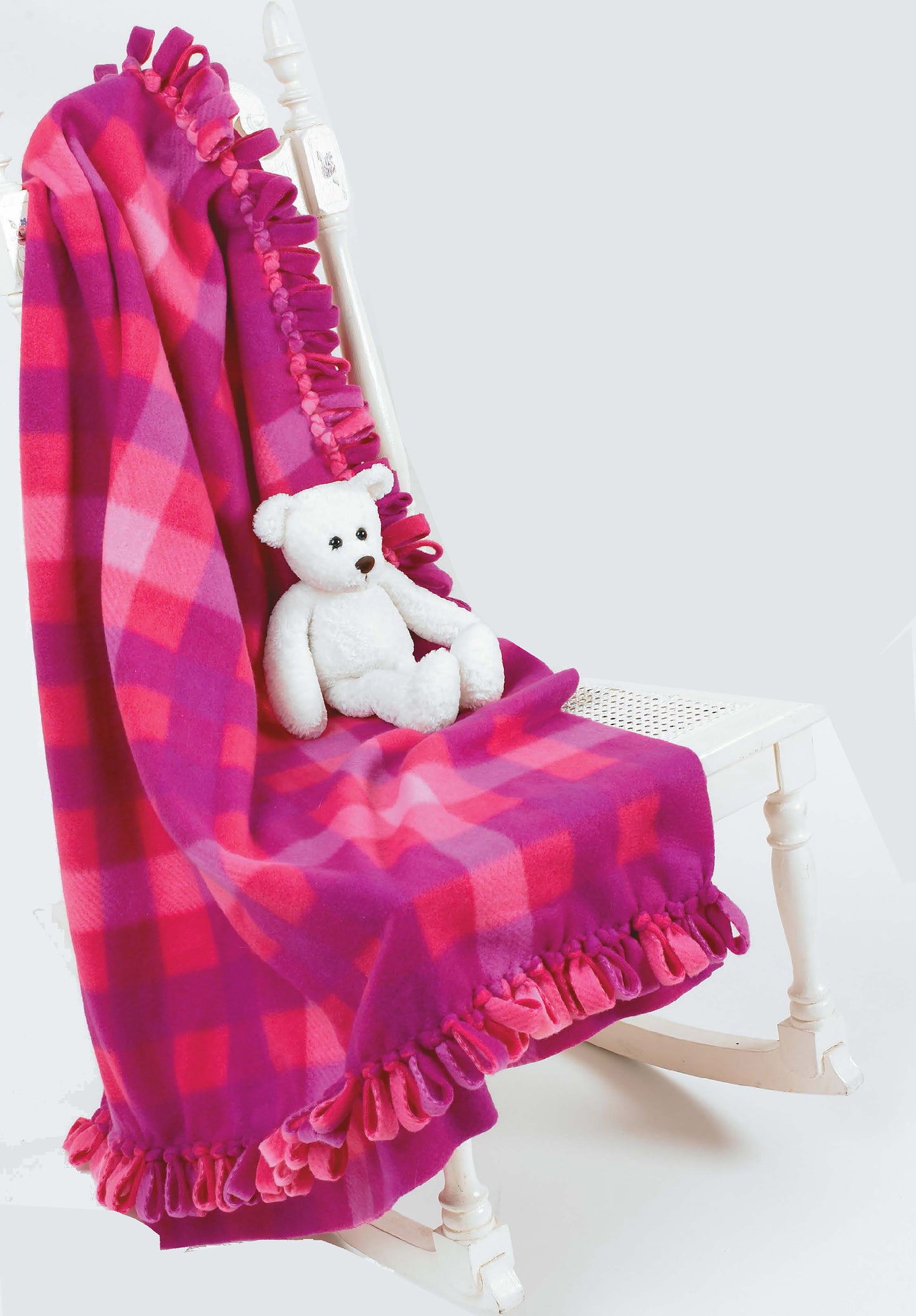 A white rocking chair displays a pink and purple plaid fleece blanket—with fringed edges from Leisure Arts' Beginner's Guide to Fleece—draped over the back, while a small white teddy bear sits on the seat.