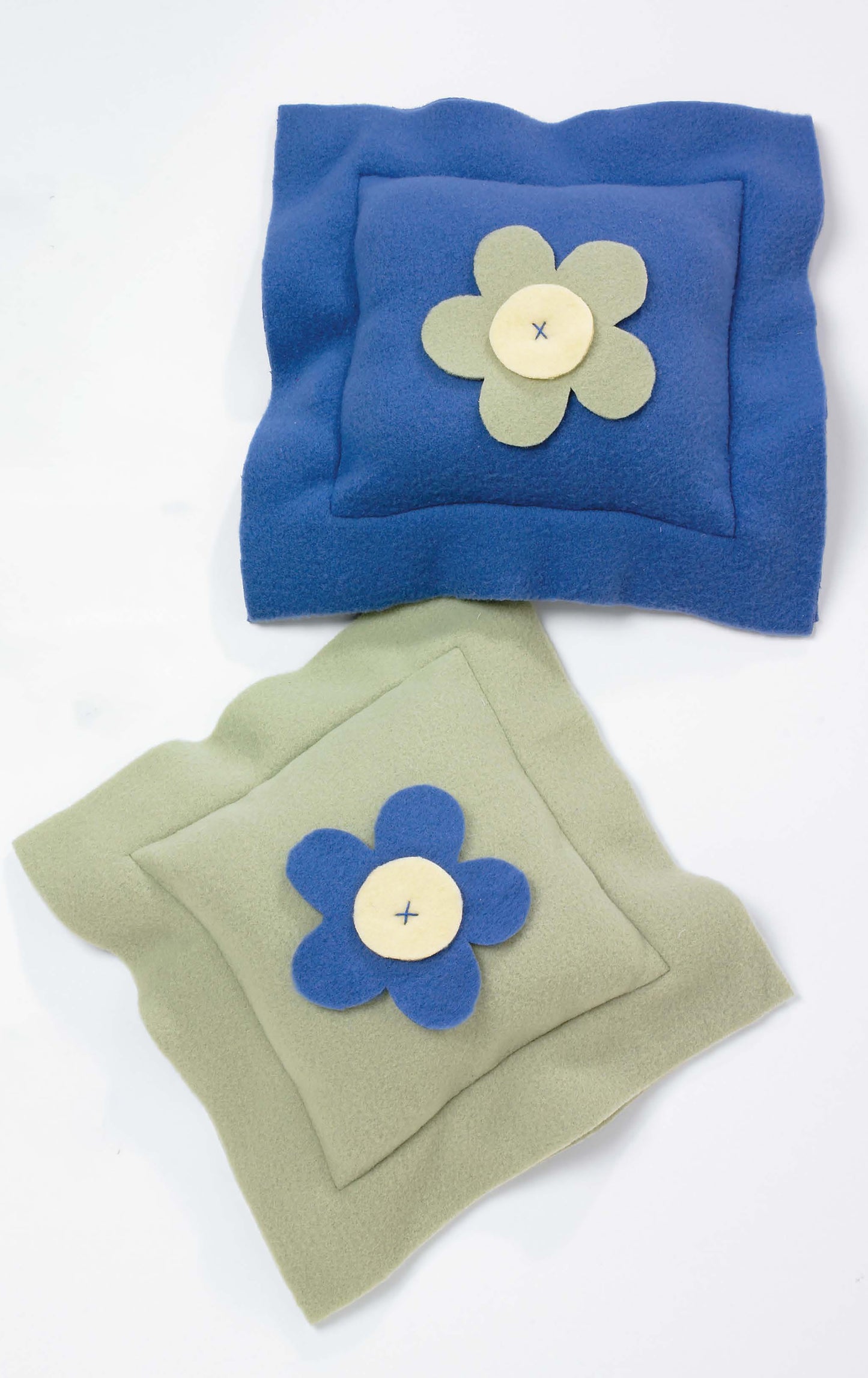 Leisure Arts' Beginner's Guide to Fleece features two square pillows—one blue with a cream and green flower, and one light green with a blue and cream flower—each with a stitched button center, displayed on a white background.