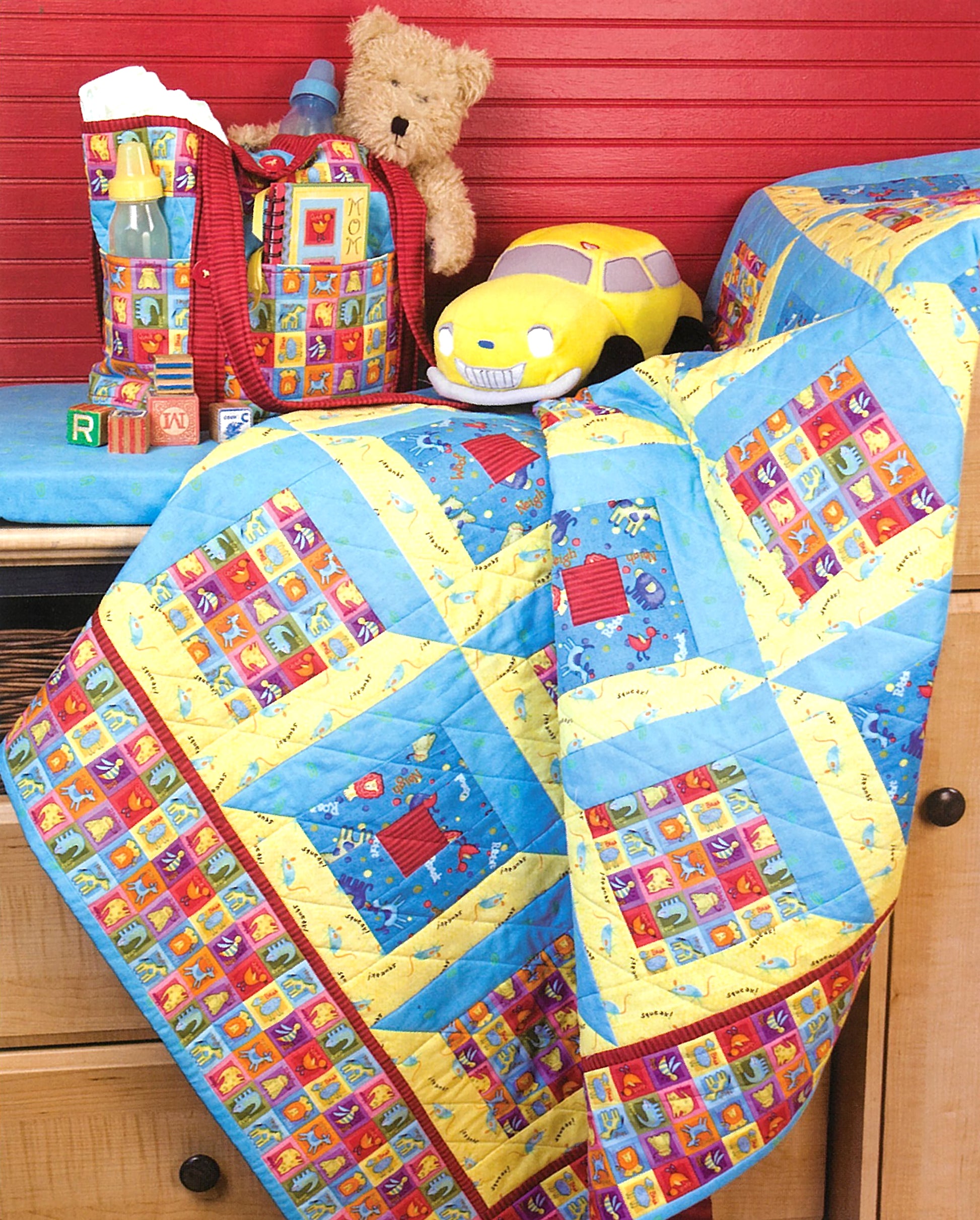 The "Debbie Mumm's Cuddle Quilts For Little Girls and Boys" by Leisure Arts, a vibrant baby quilt with blue, yellow, and patterned squares, is displayed on a dresser with baby items and toys set against a red wall.