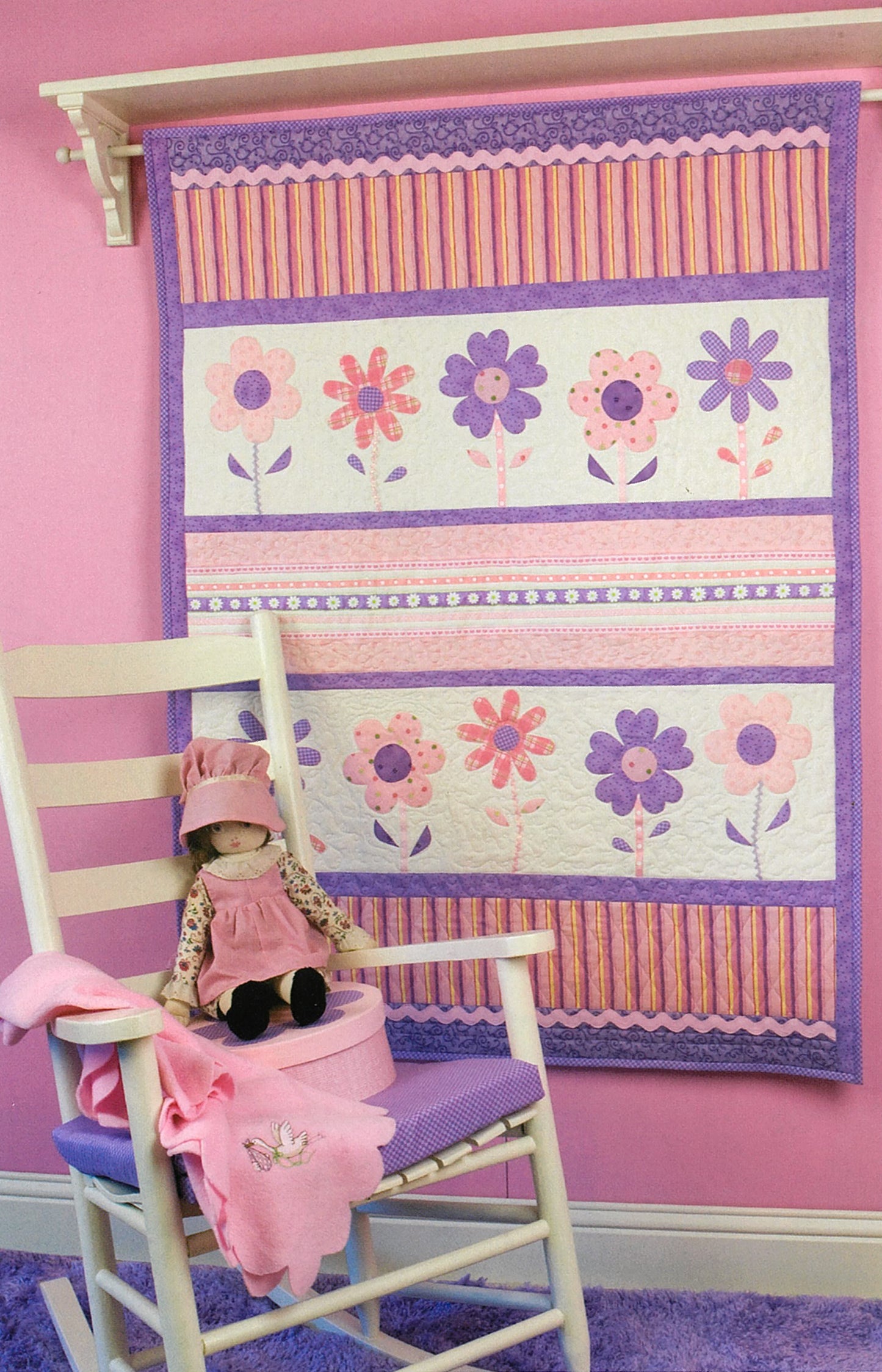 Debbie Mumm's Cuddle Quilts For Little Girls and Boys by Leisure Arts is displayed above a white rocking chair, with a doll and matching blanket in a room filled with whimsical, child-friendly decor.