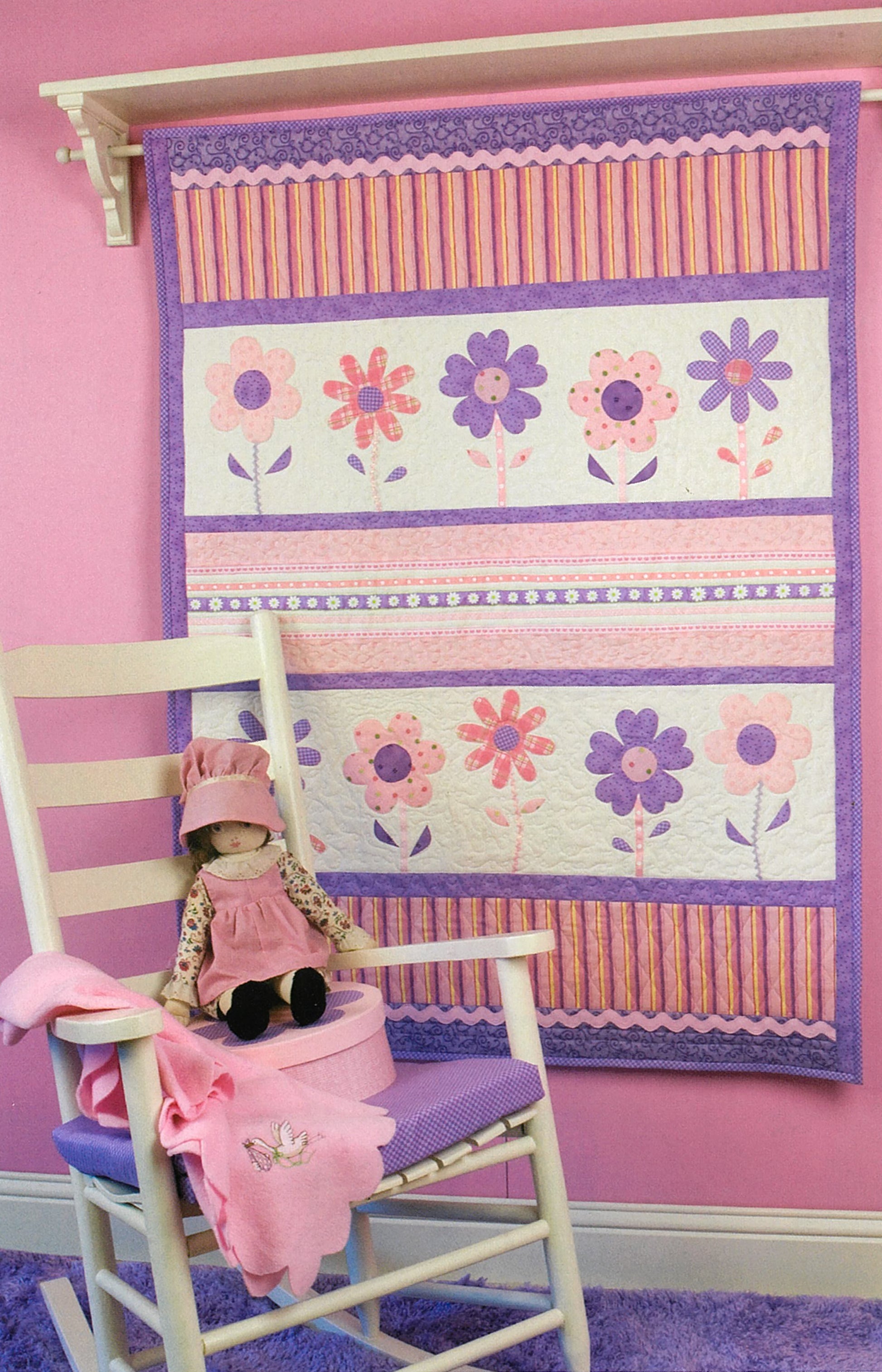 Debbie Mumm's Cuddle Quilts For Little Girls and Boys by Leisure Arts is displayed above a white rocking chair, with a doll and matching blanket in a room filled with whimsical, child-friendly decor.