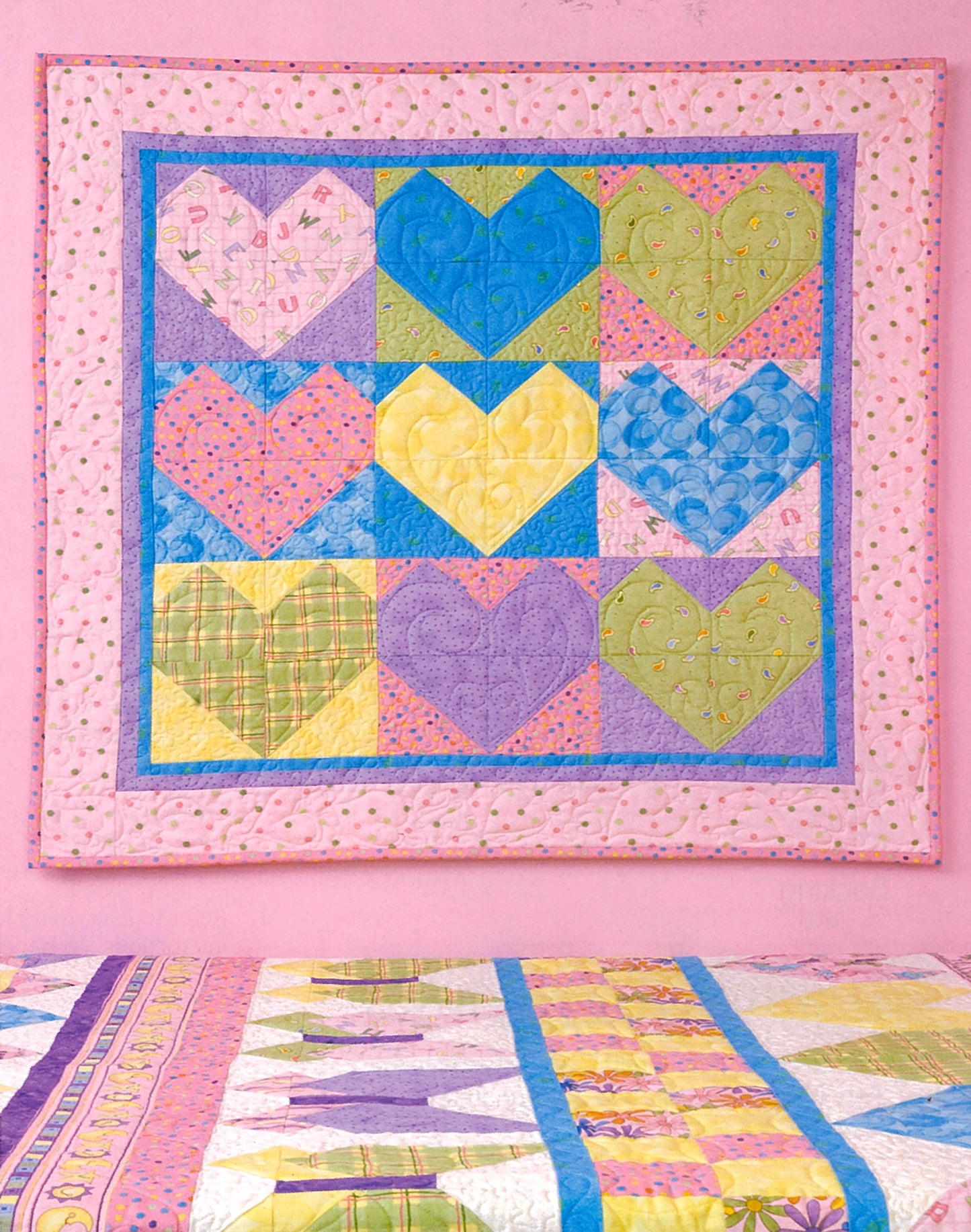A colorful quilt with twelve large, multicolored hearts hangs on a pink wall above a bed topped with Leisure Arts' "Debbie Mumm's Cuddle Quilts For Little Girls and Boys" in cheerful pastel colors.
