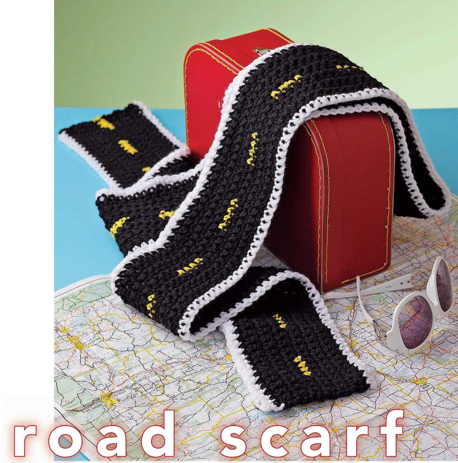 The "Crochet Your Cares Away" black worsted weight scarf by Leisure Arts features yellow dashed lines and white edging like a road. Shown over a red suitcase on a map, it’s ideal for creative crochet projects. Sunglasses and “road scarf” text appear nearby.