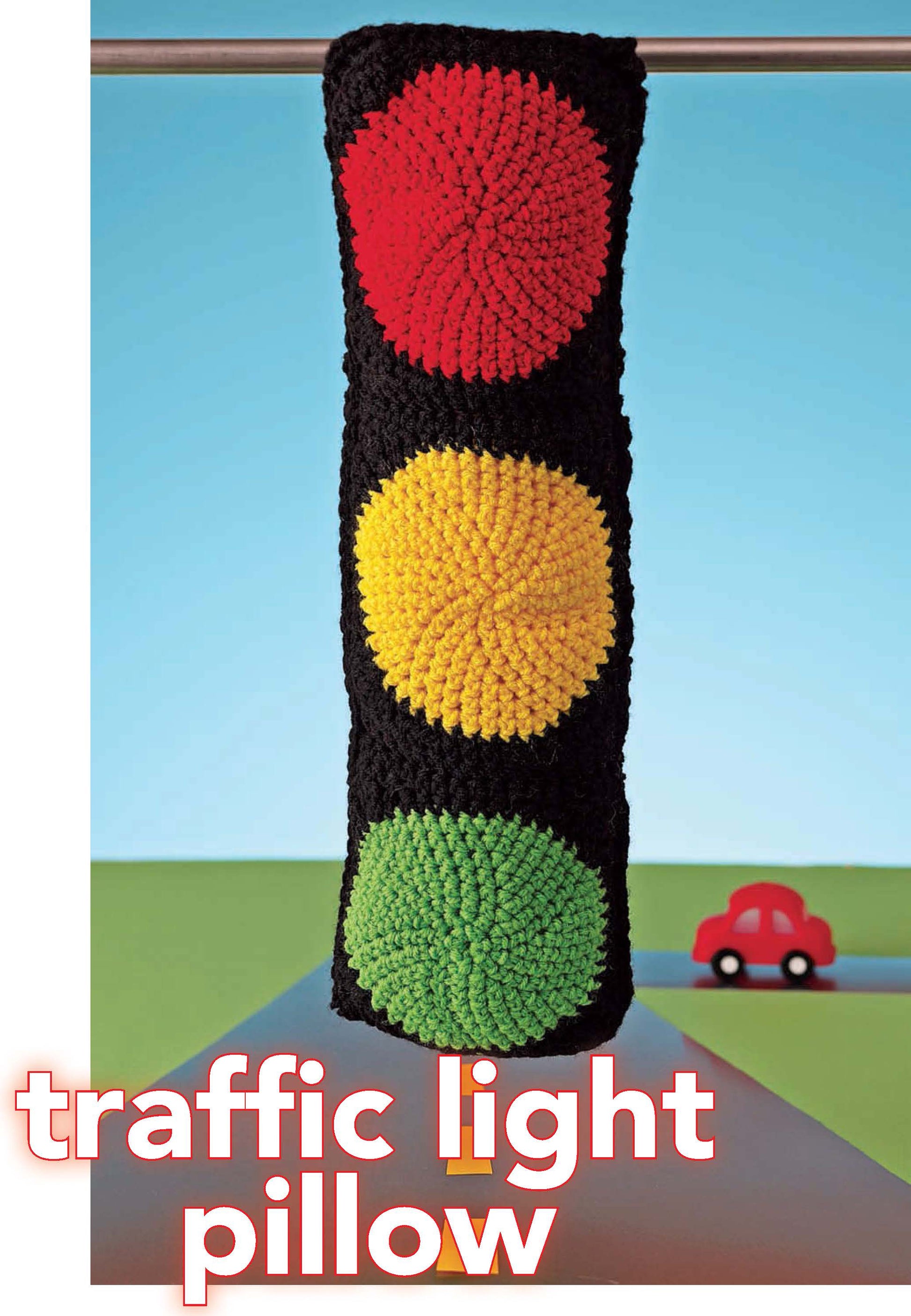 A tall, rectangular pillow from Leisure Arts’ Crochet Your Cares Away, crocheted with worsted weight yarn in red, yellow, and green like a traffic light, hangs on a rod. A small red toy car is behind it. Text reads: Crochet Your Cares Away.