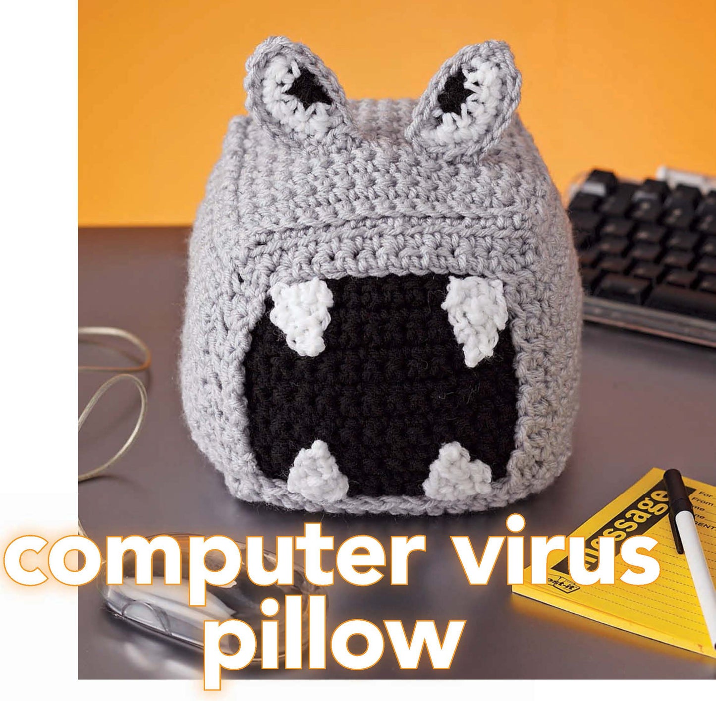 A small gray pillow, crocheted with worsted weight yarn and black and white accents, rests by a keyboard. The image text says "Crochet Your Cares Away" by Leisure Arts—perfect for fun crochet projects!.