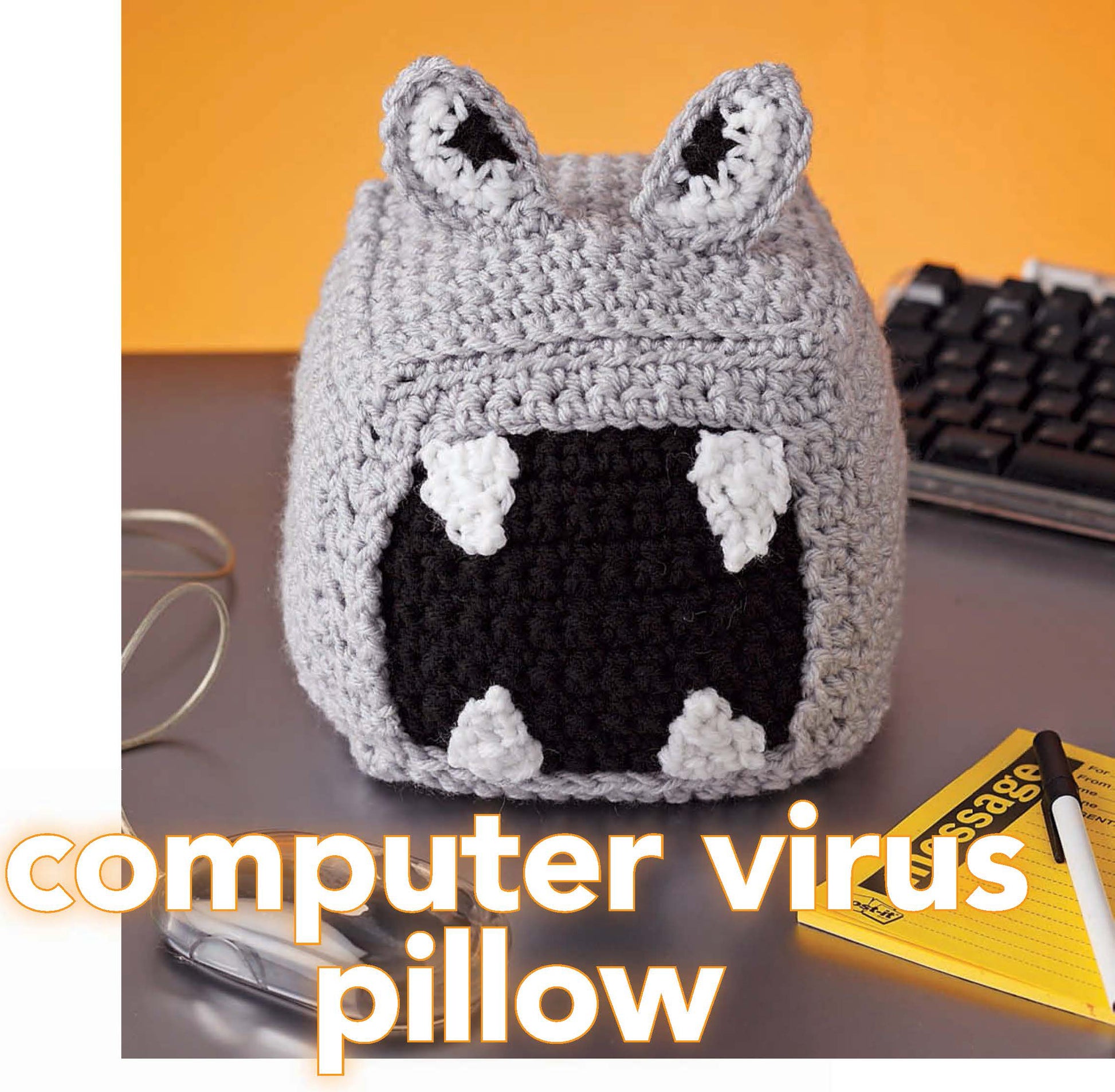 A small gray pillow, crocheted with worsted weight yarn and black and white accents, rests by a keyboard. The image text says "Crochet Your Cares Away" by Leisure Arts—perfect for fun crochet projects!.