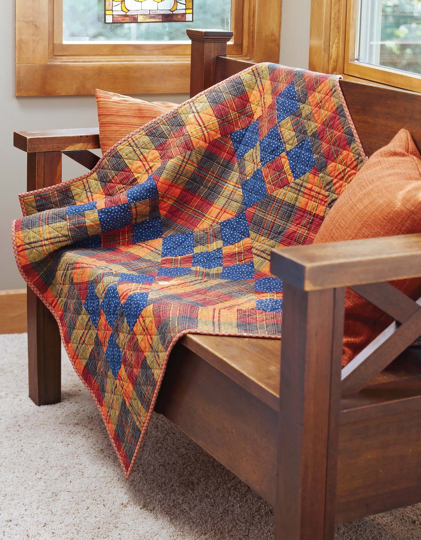 The Better Homes and Gardens 1-2-3 Quilt by Leisure Arts, featuring easy patterns and vibrant fabrics, is draped over a wooden bench with orange cushions by a sunlit window.
