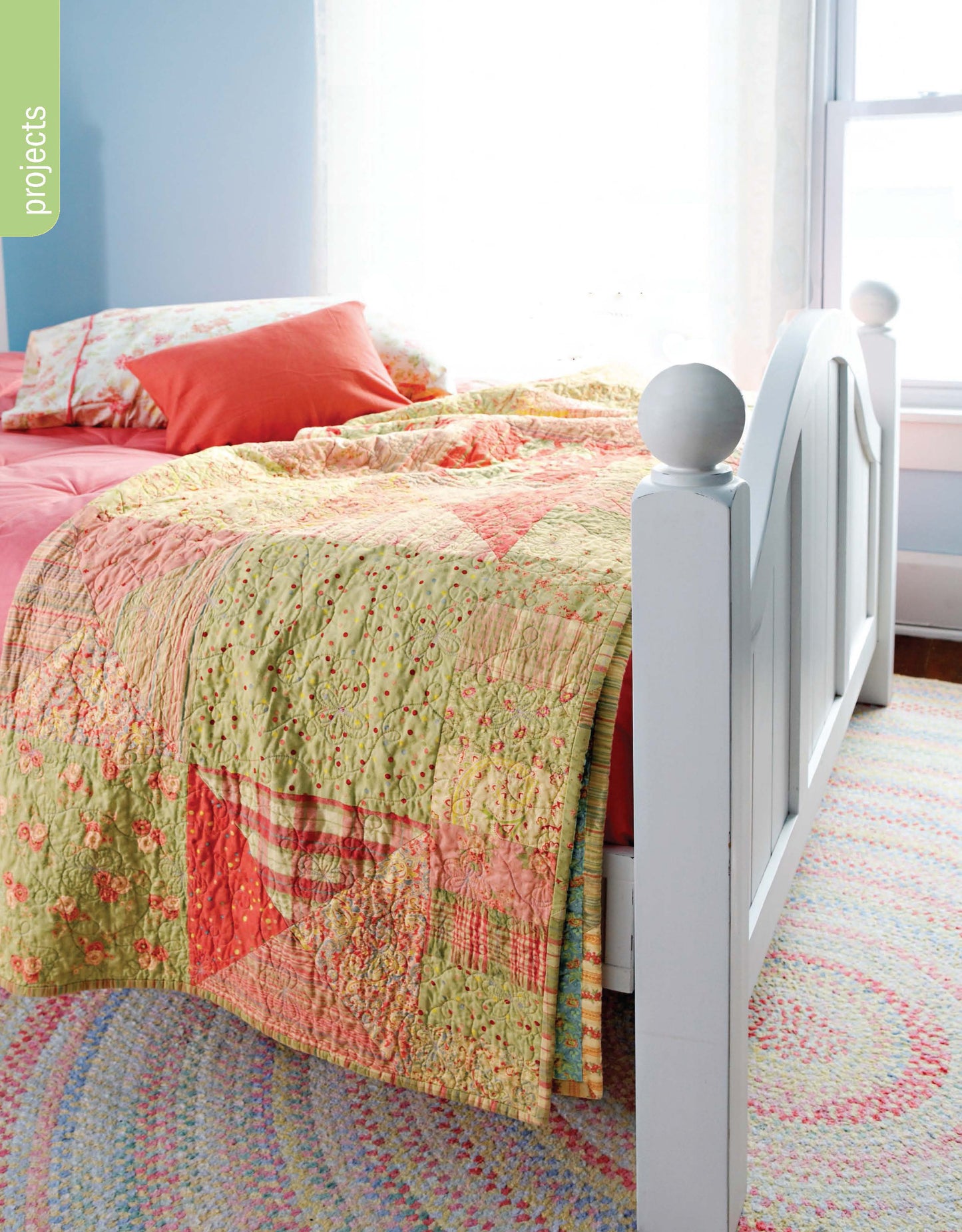 A sunlit bedroom with a white wooden bed frame, pink and orange pillows, the colorful Better Homes and Gardens 1-2-3 Quilt by Leisure Arts, and a pastel braided rug. Light streams through a window behind the bed.