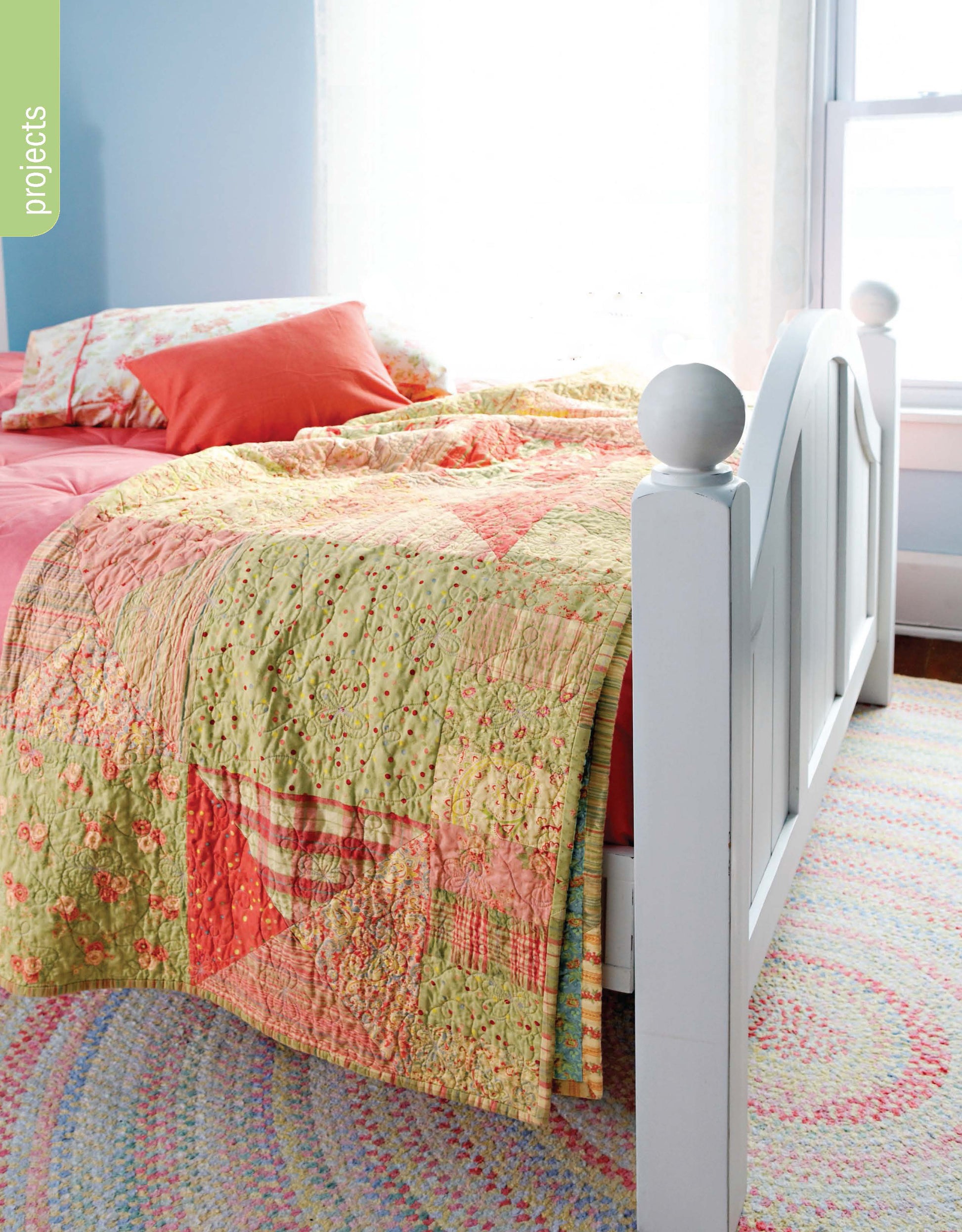 A sunlit bedroom with a white wooden bed frame, pink and orange pillows, the colorful Better Homes and Gardens 1-2-3 Quilt by Leisure Arts, and a pastel braided rug. Light streams through a window behind the bed.