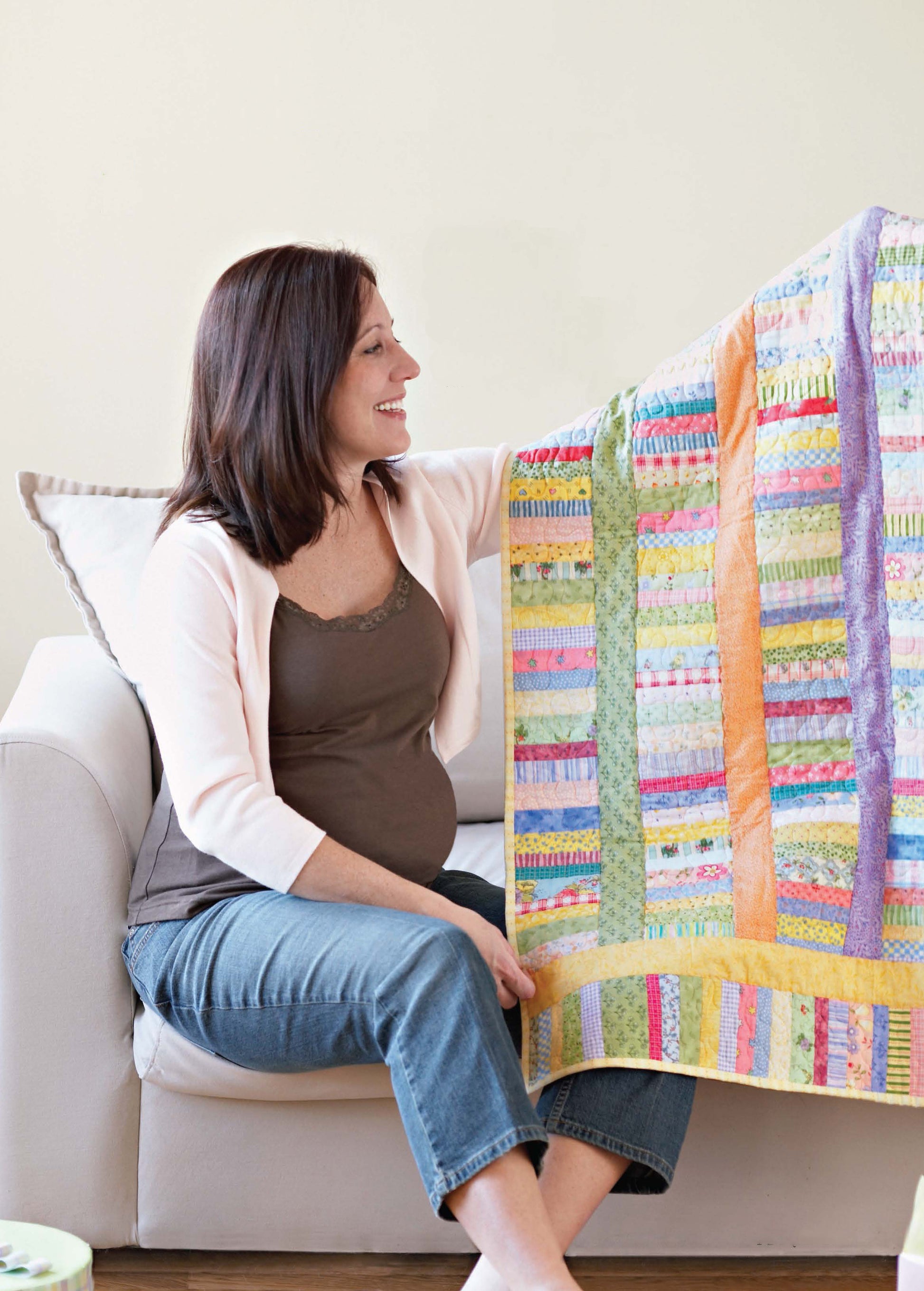 A woman sits on a sofa, smiling and holding up a colorful quilt made from Better Homes and Gardens 1-2-3 Quilt by Leisure Arts—an inspiring project featuring easy patterns perfect for beginners.