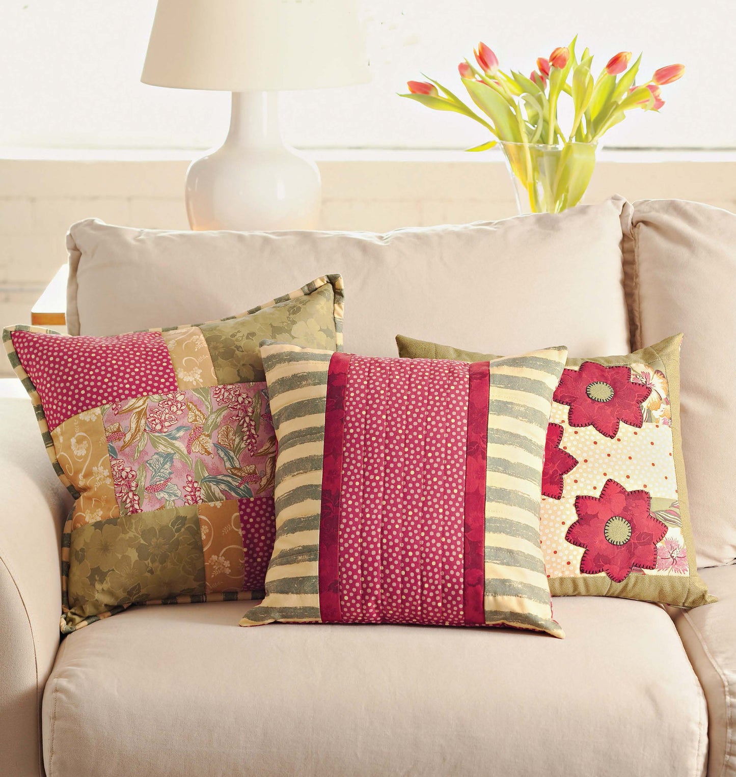 Three colorful patchwork pillows, made with the Better Homes and Gardens 1-2-3 Quilt by Leisure Arts, are displayed on a beige sofa. Behind them, a white lamp and tulips in a vase add charming touches to the scene.