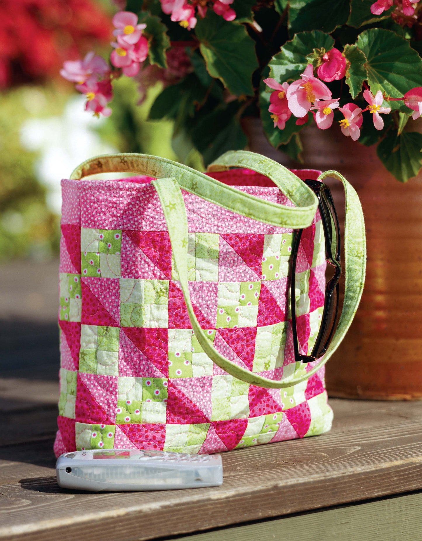 The Better Homes and Gardens 1-2-3 Quilt by Leisure Arts—a vibrant pink, green, and white patchwork tote for beginner quilters—rests on wood beside a glasses case, with a flowering plant in the background.
