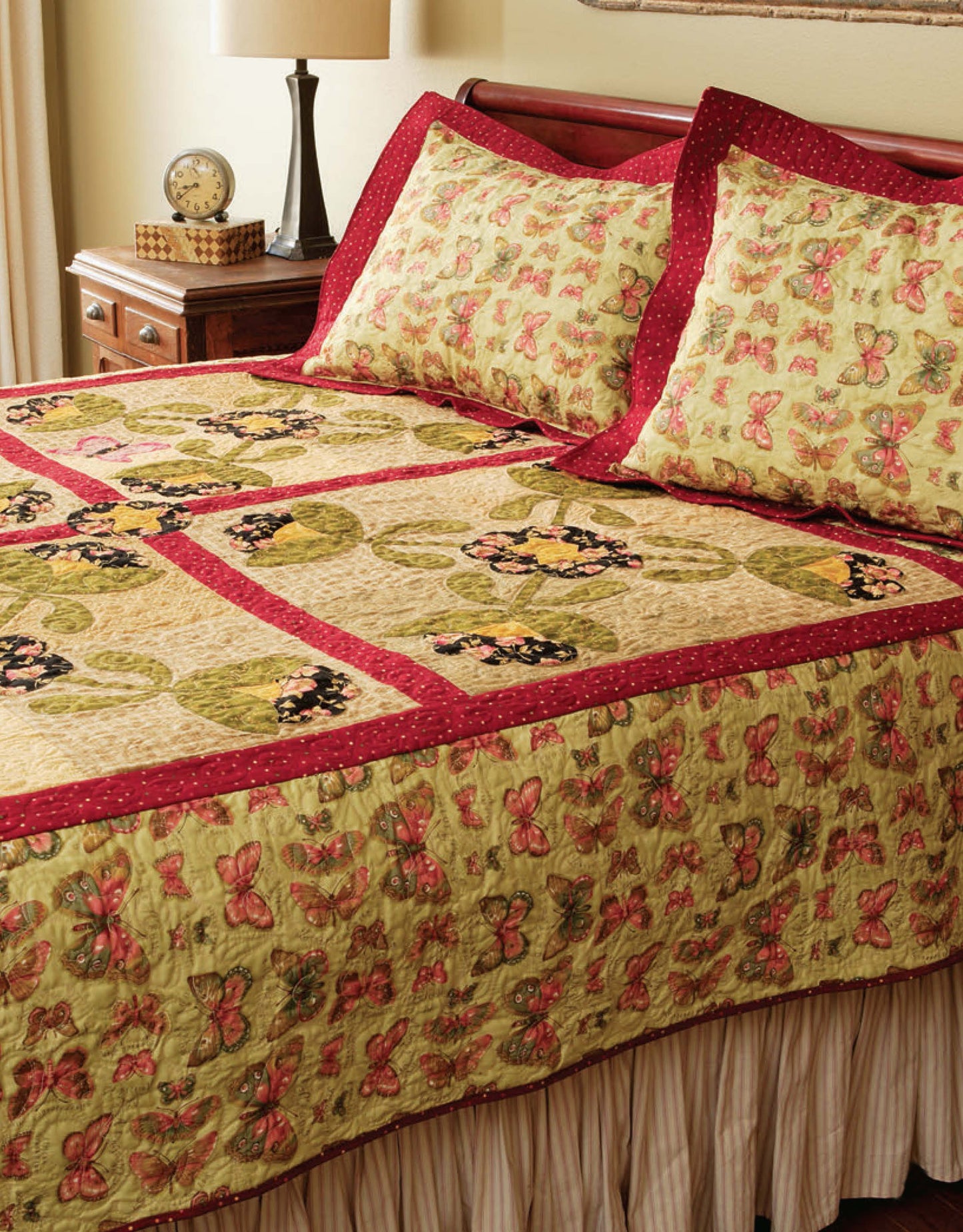 A neatly made bed features a butterfly and leaf quilt in green, yellow, pink, and red—perfect inspiration for beginner projects using "Learn to Machine Quilt with Pat Sloan" by Leisure Arts. A nightstand with a lamp, clock, and book sits nearby.