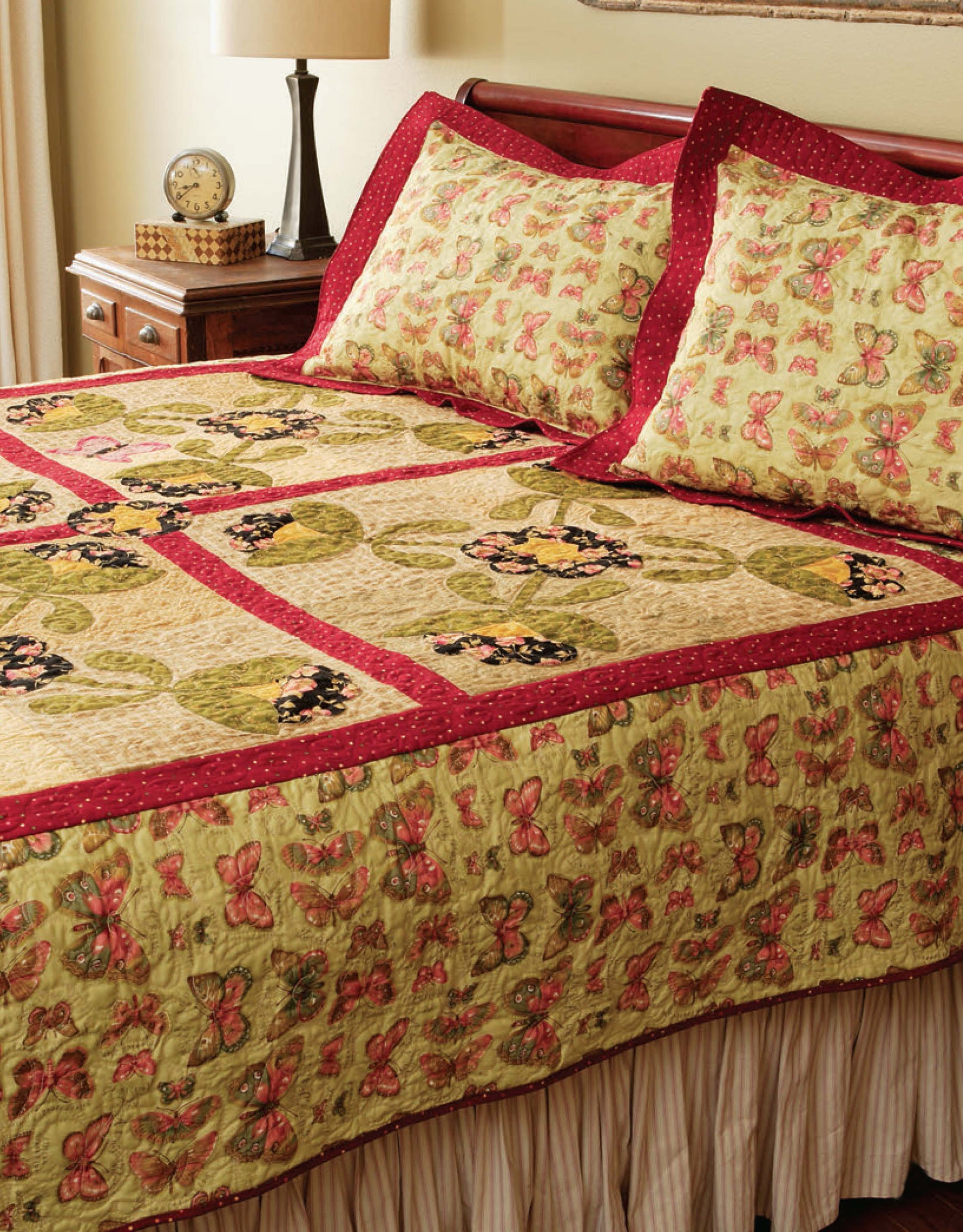 A neatly made bed features a butterfly and leaf quilt in green, yellow, pink, and red—perfect inspiration for beginner projects using "Learn to Machine Quilt with Pat Sloan" by Leisure Arts. A nightstand with a lamp, clock, and book sits nearby.