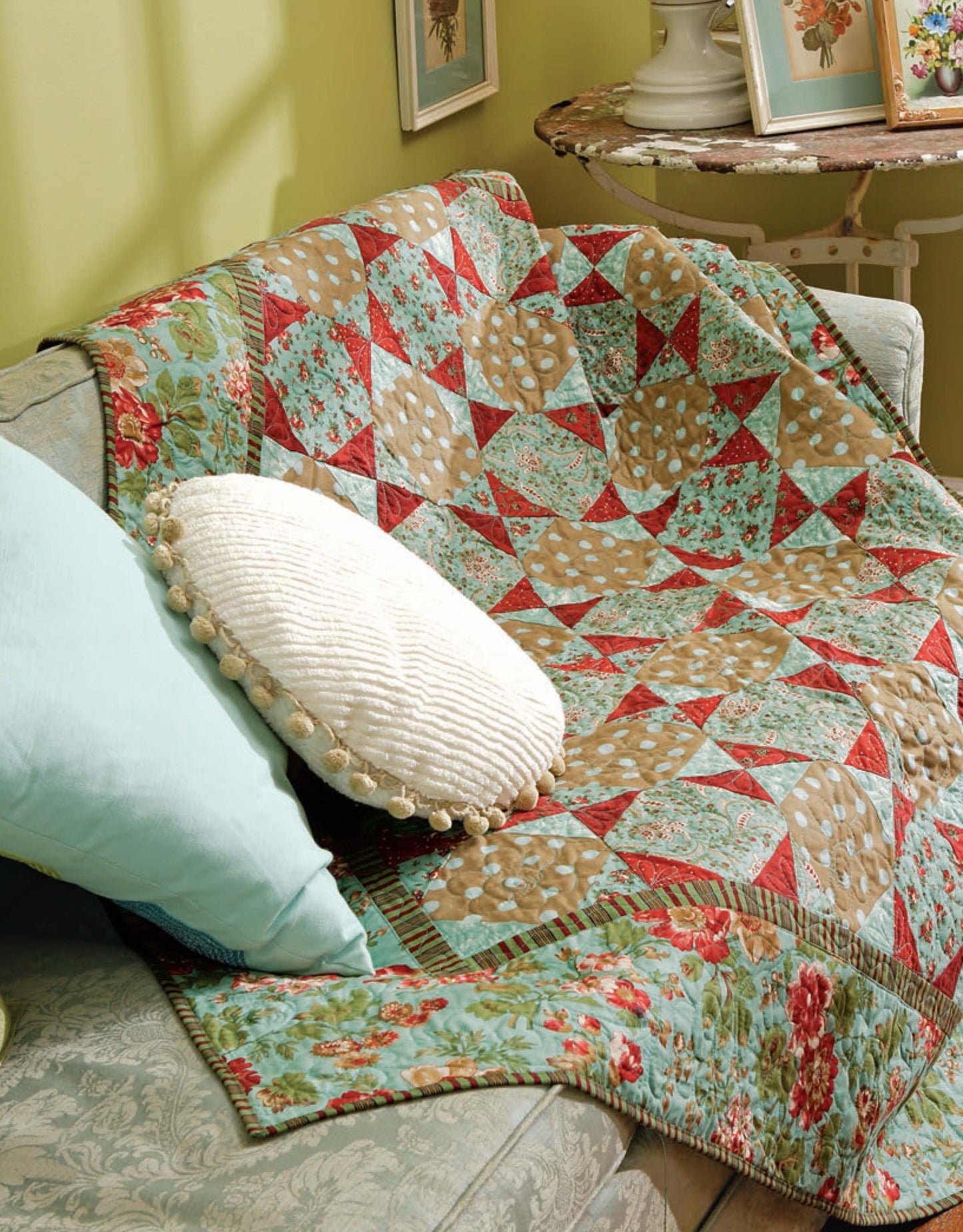 A colorful quilt inspired by "Learn to Machine Quilt with Pat Sloan" by Leisure Arts drapes over a light blue sofa with a round white pillow. Framed pictures and a small side table appear in the background.