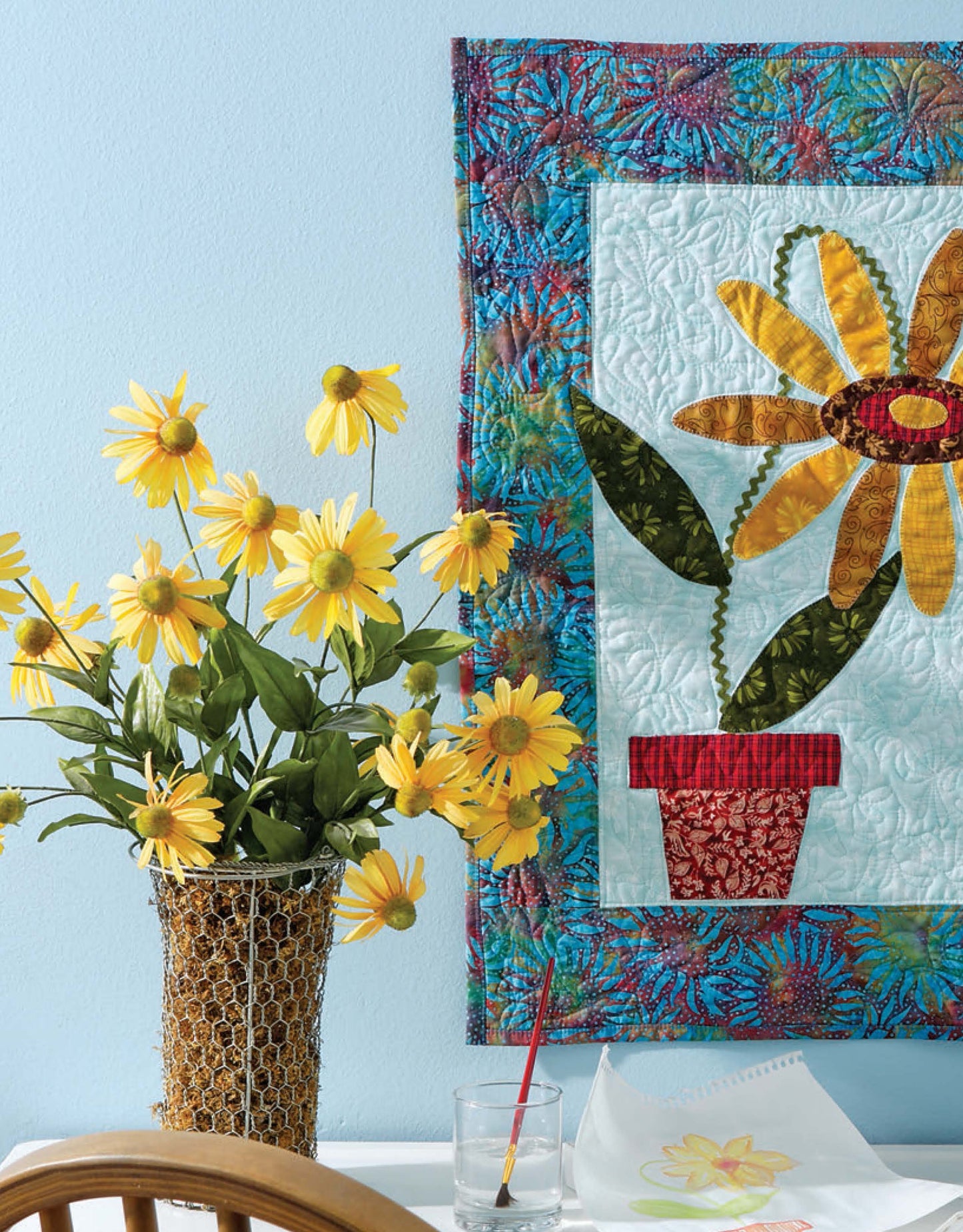 A vase of yellow flowers sits on a table with a glass of water, a paintbrush, and a flower sketch. Behind them hangs "Learn to Machine Quilt with Pat Sloan" by Leisure Arts—a colorful quilt perfect for beginner quilting projects.
