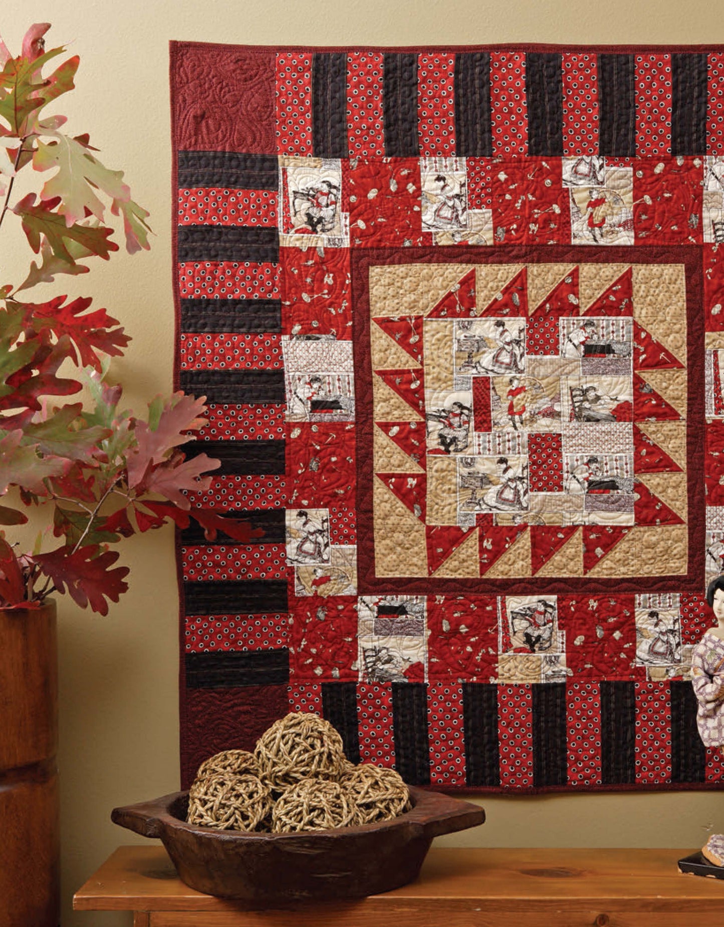 A vibrant quilt inspired by "Learn to Machine Quilt with Pat Sloan" from Leisure Arts hangs on the wall, while a table nearby displays wicker balls in a bowl and a vase with leafy branches—perfect for beginner quilting inspiration.