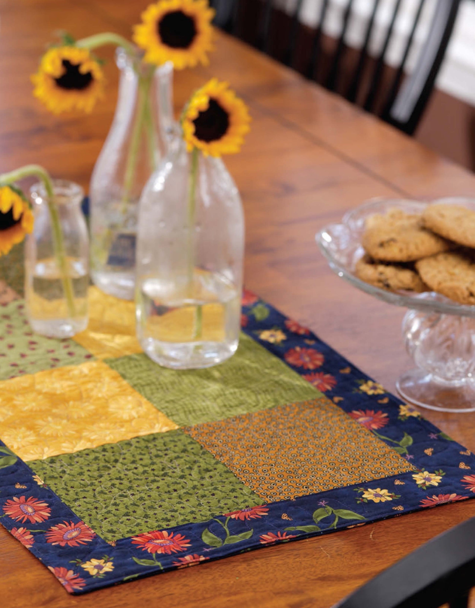Three glass vases with sunflowers and water sit on a colorful quilted table runner inspired by Leisure Arts’ “Learn to Machine Quilt with Pat Sloan,” next to a glass pedestal plate of cookies on a wooden dining table.