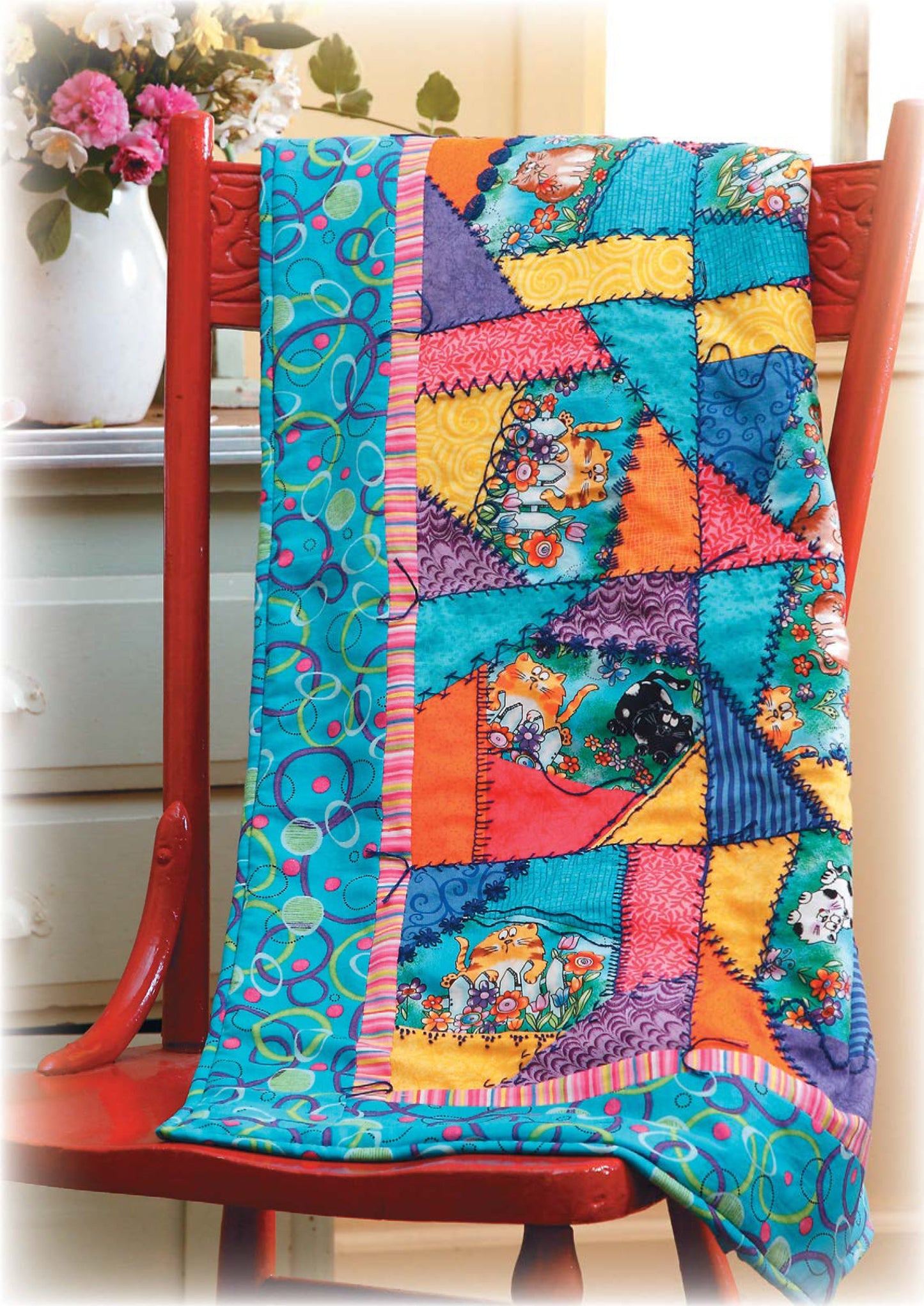 The Quilt Along with Emilie Richards by Leisure Arts, featuring vibrant traditional patterns, is draped over a red wooden chair beside a flower vase and light green dresser, adding cozy charm to the scene.