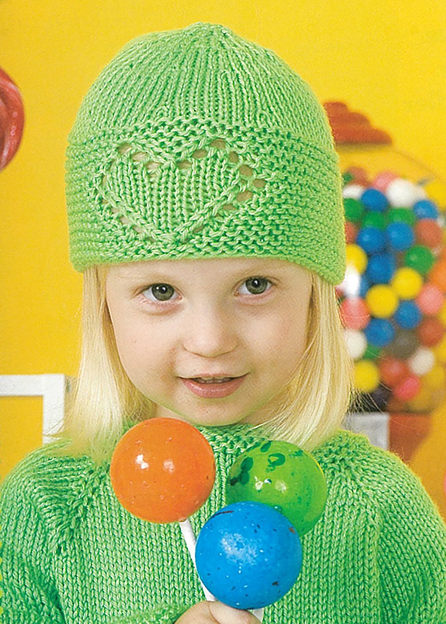 A young blond child wearing a green hat and sweater from "Lots of Love Knits for Kids" by Leisure Arts holds three large lollipops, with a gumball machine in the bright yellow background.