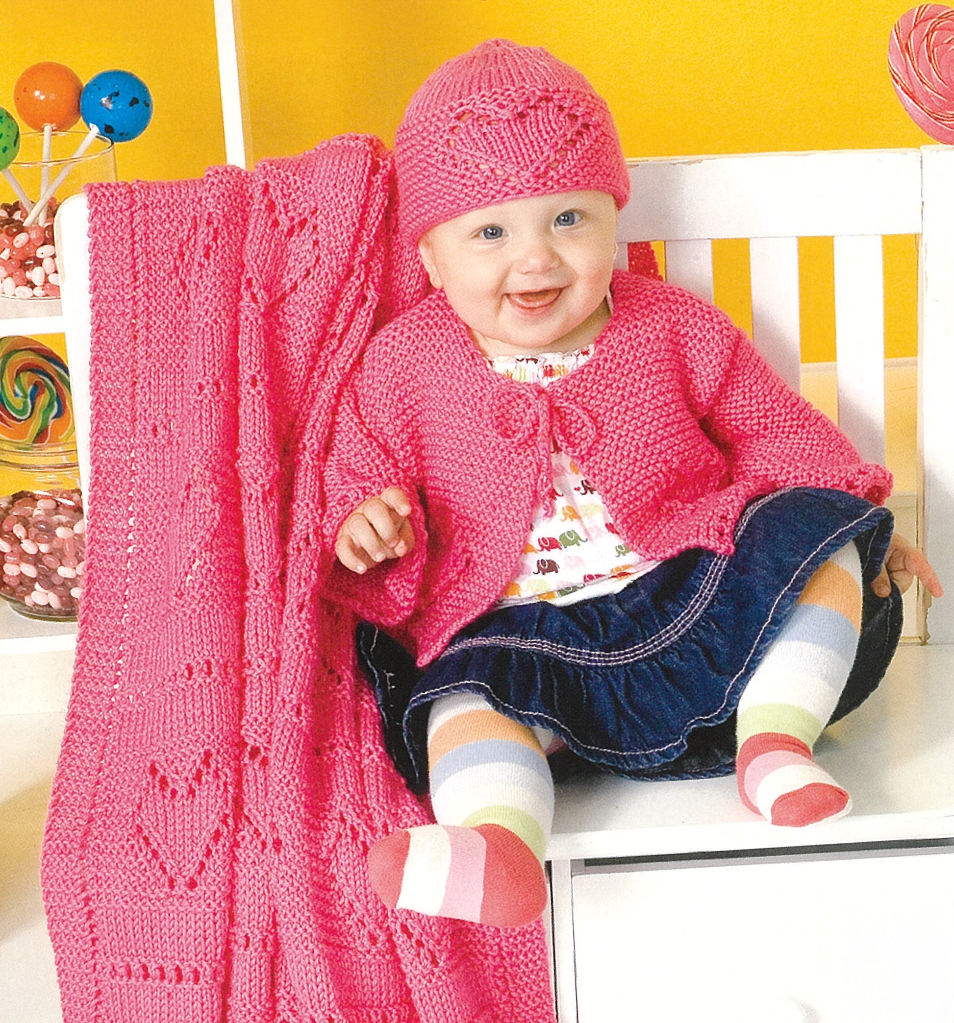 A baby in a pink knit hat and sweater from Leisure Arts' "Lots of Love Knits for Kids" sits on a white bench with a denim skirt and striped tights. A pink heart-patterned knitted blanket is beside the baby, with colorful candy jars in the background.