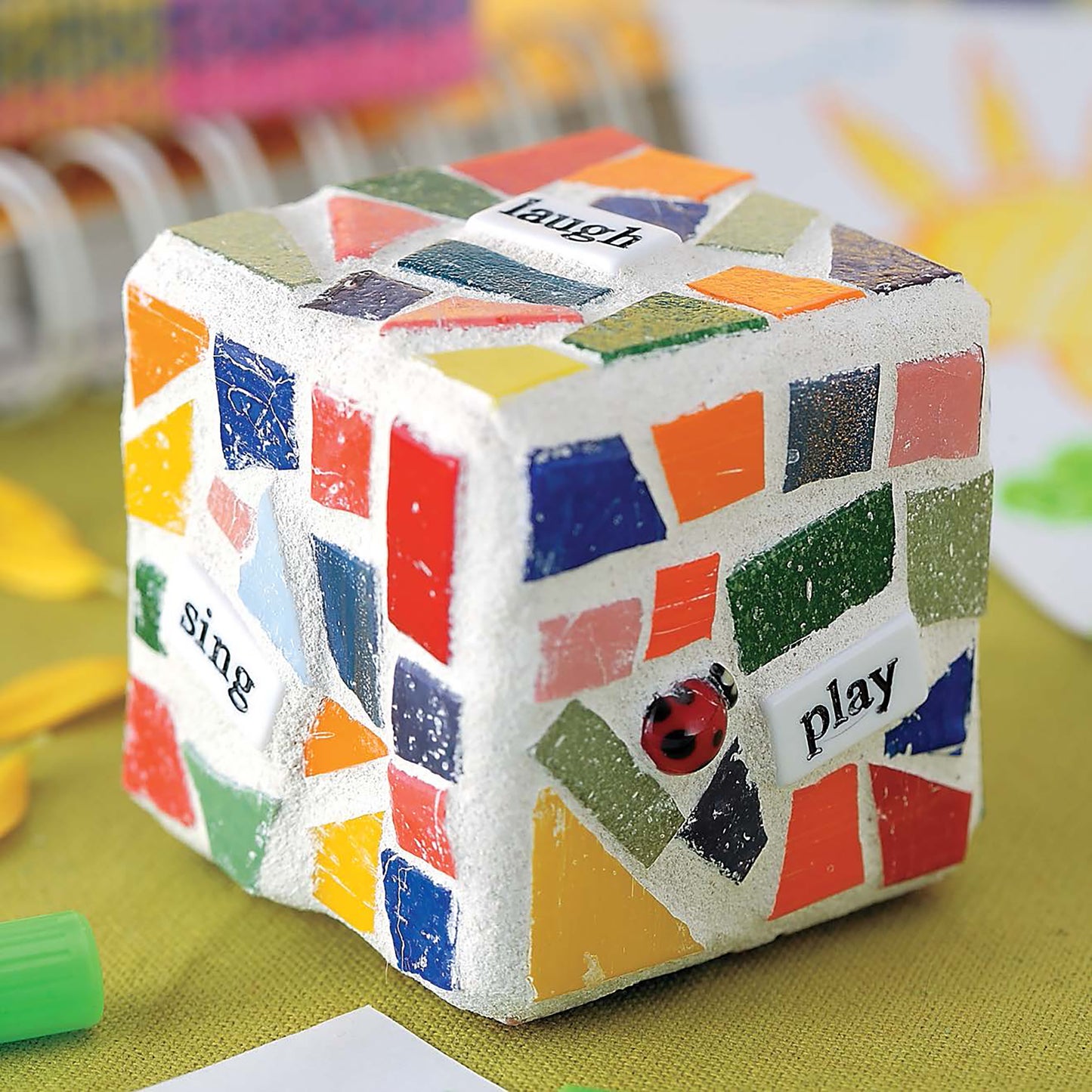 Heidi Borchers' Beginner's Guide to Mosaics by Leisure Arts features a white cube with vibrant tile inlays, words like sing, laugh, play on its sides, and a ladybug accent—perfect for adding charm to your home decor.