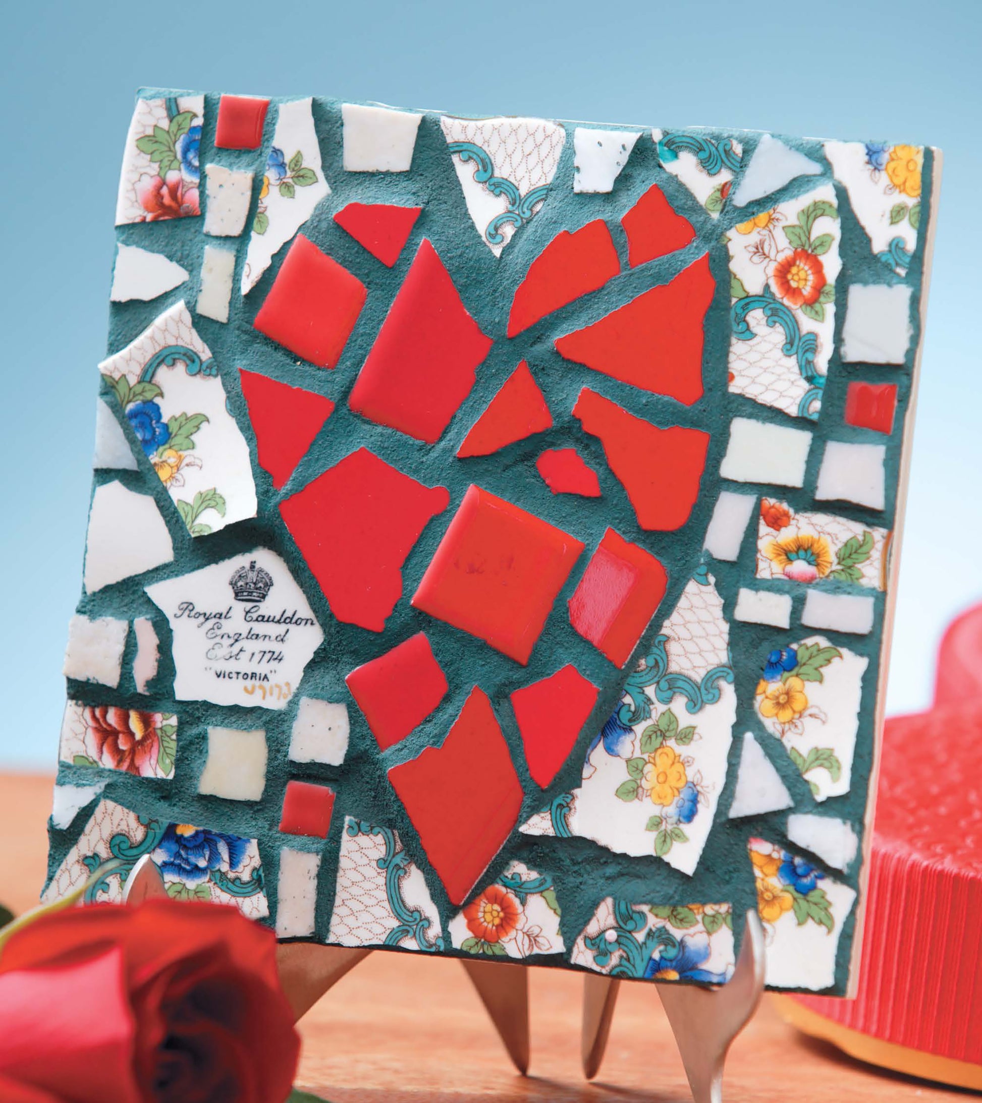 Heidi Borchers' Beginner's Guide to Mosaics by Leisure Arts shows how to create mosaic art, like a red broken heart design with tile and floral china on a square base, displayed on an easel with a red rose in front.