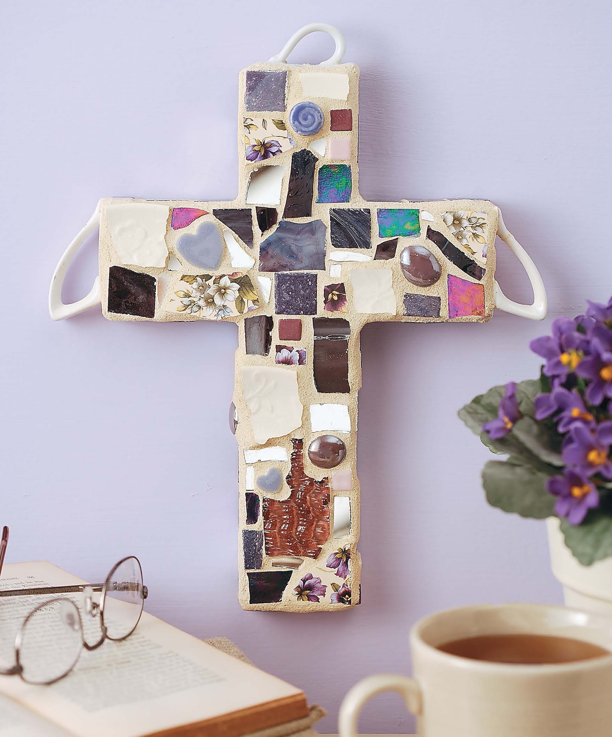 Heidi Borchers' Beginner's Guide to Mosaics by Leisure Arts rests beside purple flowers, glasses, and coffee on a table, while a colorful mosaic cross decorates a light purple wall for cozy home decor.