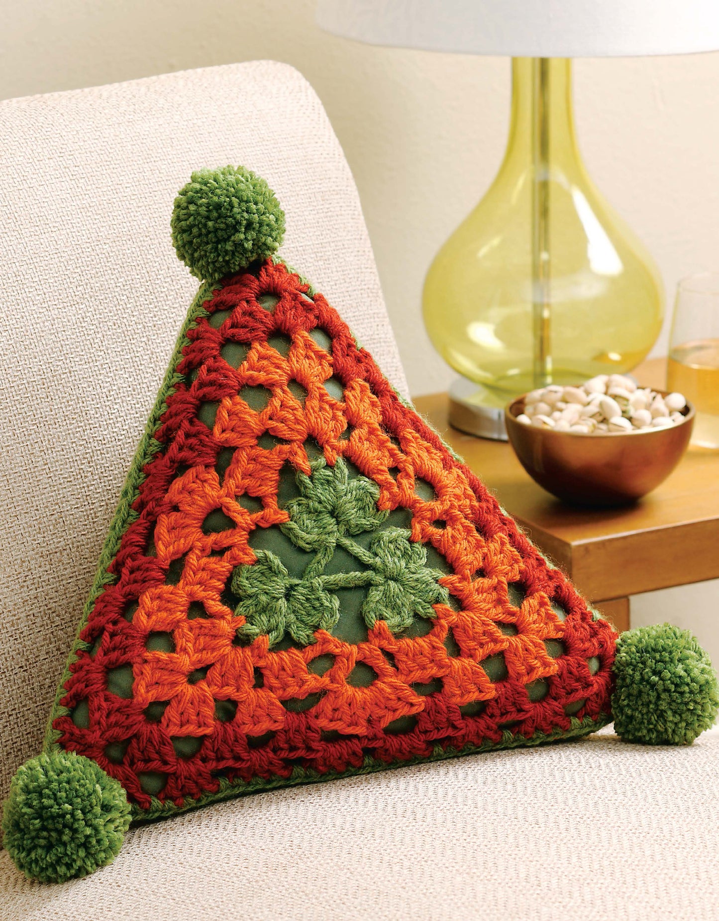The Triangle Treasury crochet pillow by Leisure Arts features orange, red, and green triangle patterns with three green yarn pom-poms on its corners. It sits on a beige chair beside a table with a lamp and bowl of snacks.