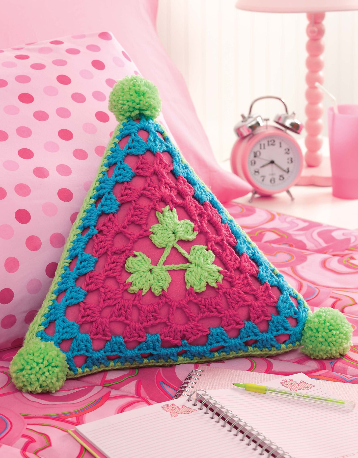 A vibrant pillow crafted from Triangle Treasury motifs by Leisure Arts, finished with green corner pom-poms, rests on a pink polka-dot bed; nearby are a pink clock, lamp, and a notepad with pen.