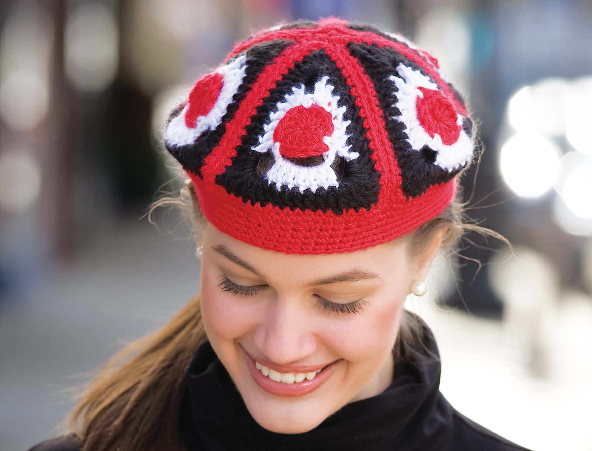 A woman smiles and looks down, wearing a vibrant hat made from Leisure Arts' Triangle Treasury—A Must-Have for Your Crochet Library! The bold red, black, and white triangle motifs stand out against the blurred background.