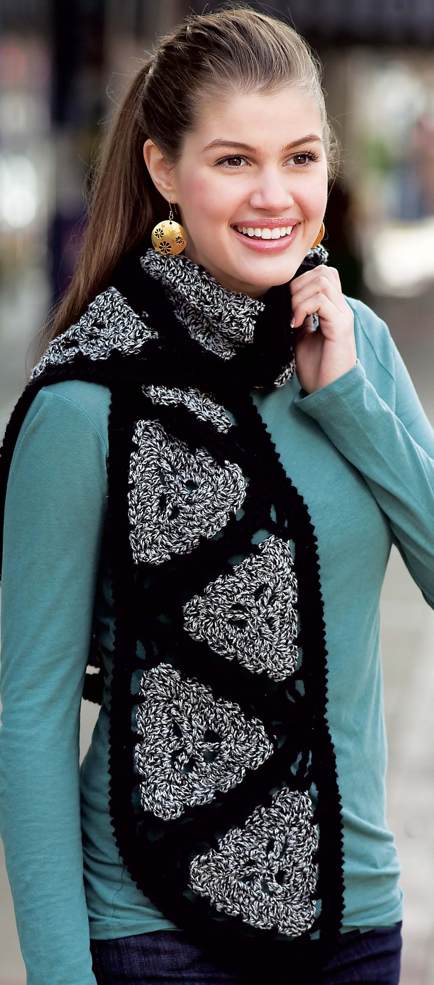 A smiling woman with long brown hair in a ponytail wears a teal long-sleeve shirt and showcases the Leisure Arts Triangle Treasury scarf, highlighting creative crochet projects for your library.