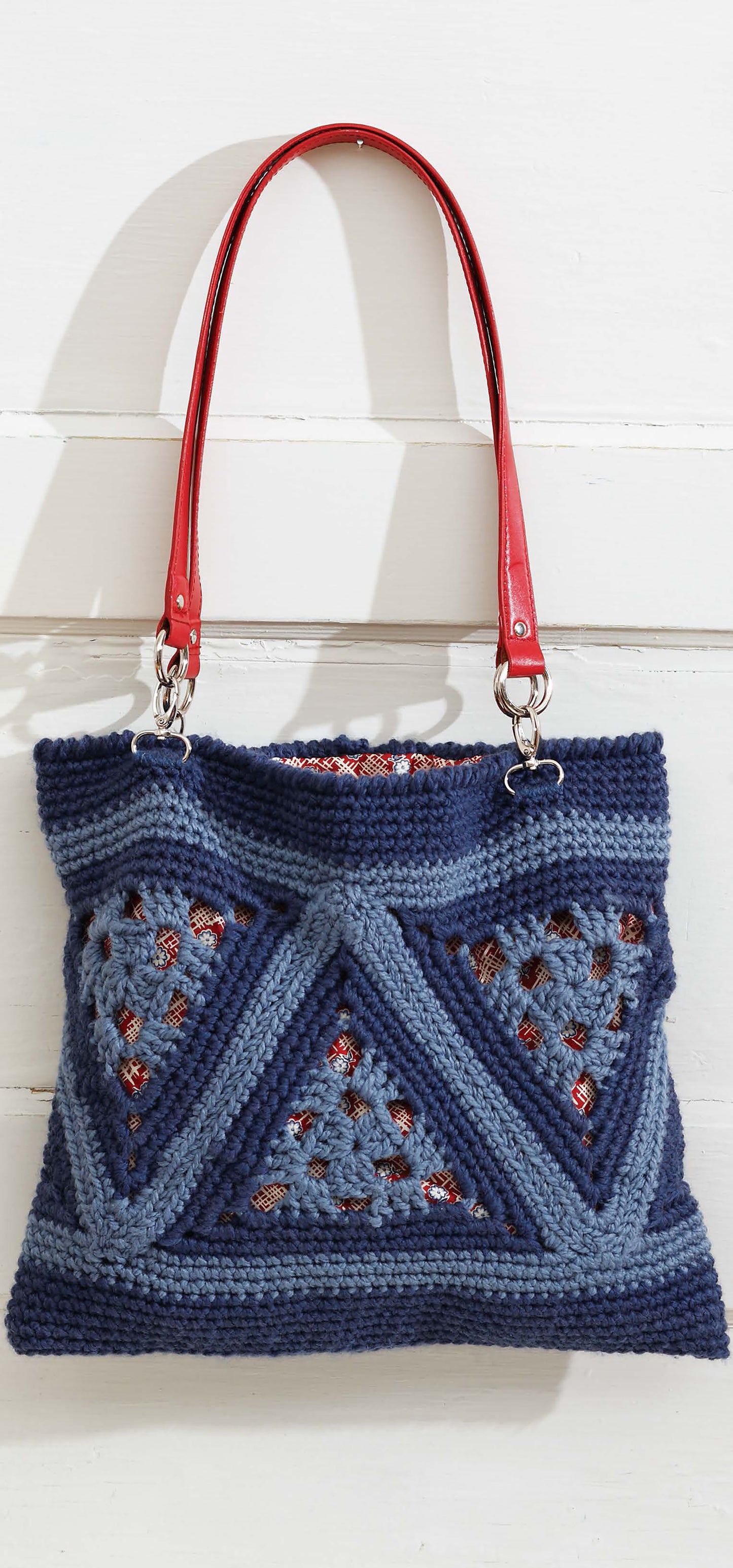 Triangle Treasury by Leisure Arts, a stylish blue crocheted handbag with triangle motifs and geometric patterns, red leather handles, and silver hardware, is displayed against a white wooden background.