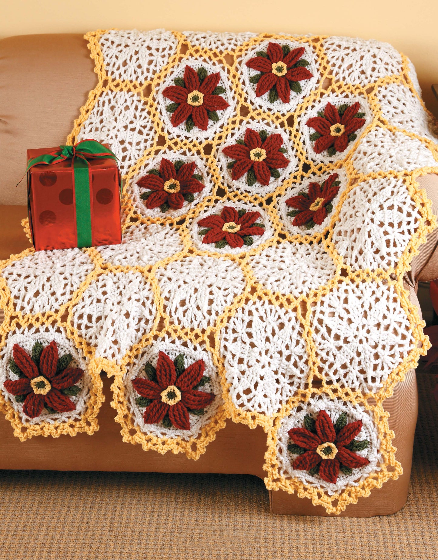 Floral Touches Digital Download by Leisure Arts features a crocheted afghan with white and yellow hexagons, each with a red and brown flower, draped on a tan couch—a lovely choice for garden-themed crochet fans. A red gift box with green ribbon is nearby.
