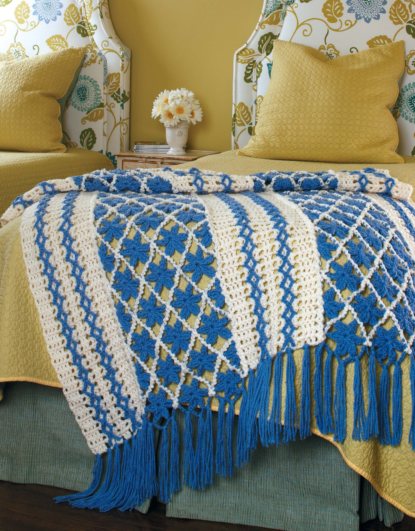 A bed is adorned with Leisure Arts' Floral Touches Digital Download crochet afghan, featuring blue flowers, white stripes, and blue fringe. Two yellow pillows and a blue-green bedspread complete the garden-themed look; white flowers sit on the bedside table.