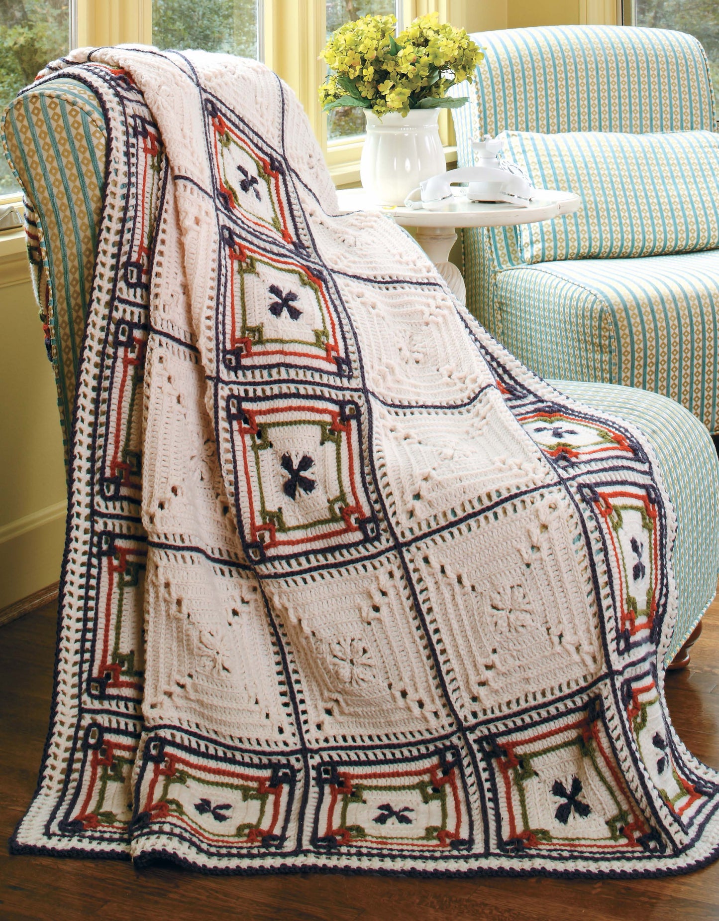 The Floral Touches Digital Download by Leisure Arts features an intermediate crochet pattern with a geometric square design in white, navy, red, and green—perfect for draping over your favorite chair as shown beside yellow flowers in a vase.