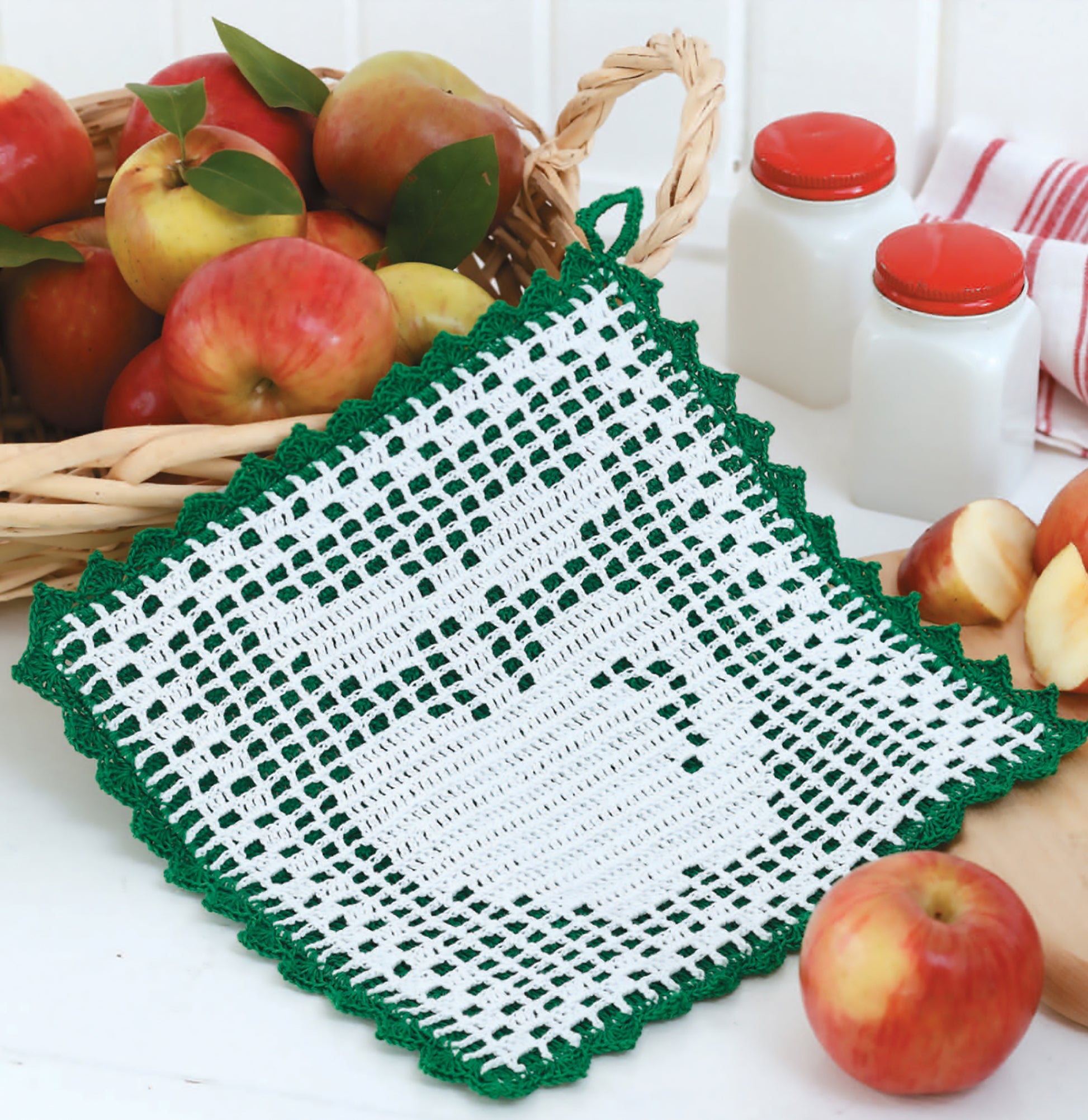 The Gooseberry Patch Farmhouse Crochet Digital Download by Leisure Arts features a white doily with a green border and apple design, shown on a table with red apples, a basket, milk bottles, and sliced apples. Perfect for beginner crochet projects.