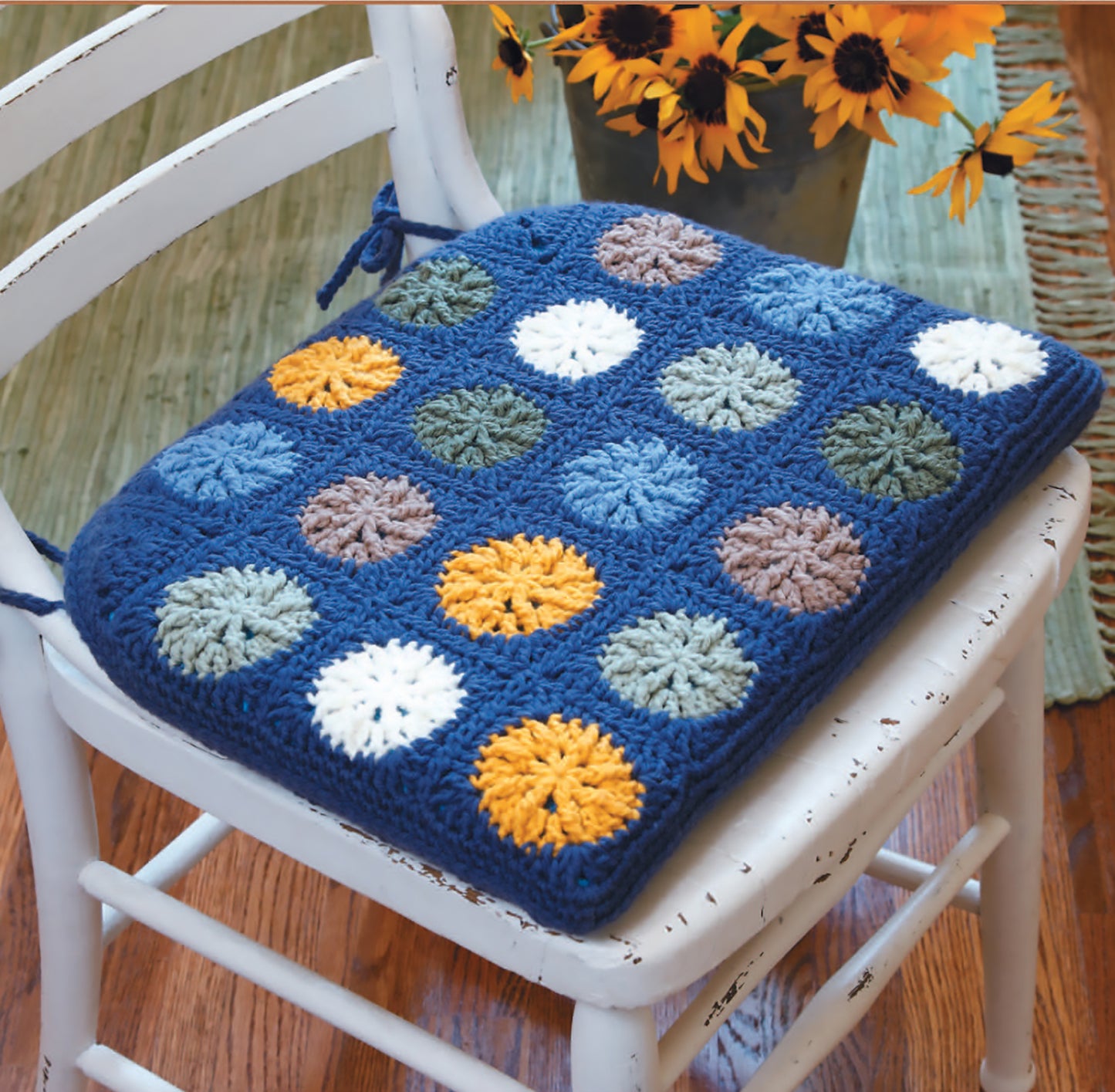 A white wooden chair with a blue seat cushion made using the Gooseberry Patch Farmhouse Crochet Digital Download by Leisure Arts, featuring colorful circular patterns, sits beside a sunflower vase on a wooden floor.