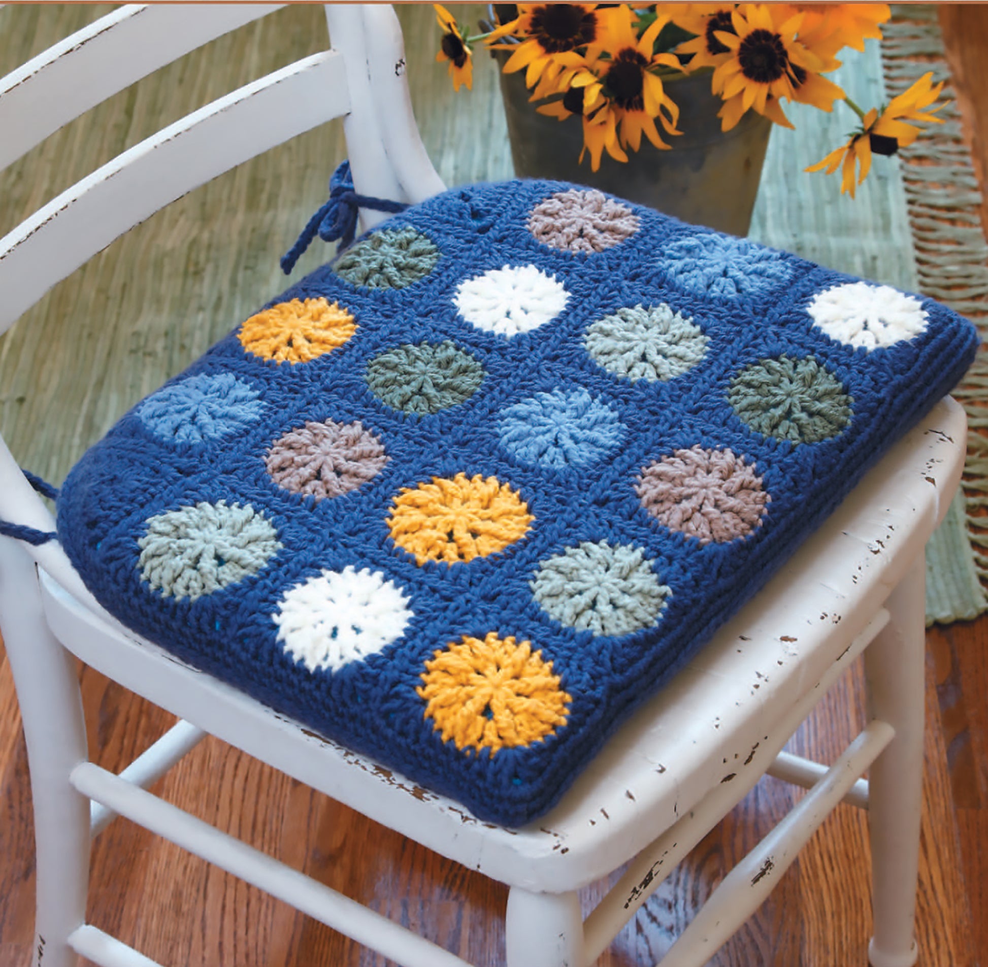 A white wooden chair with a blue seat cushion made using the Gooseberry Patch Farmhouse Crochet Digital Download by Leisure Arts, featuring colorful circular patterns, sits beside a sunflower vase on a wooden floor.