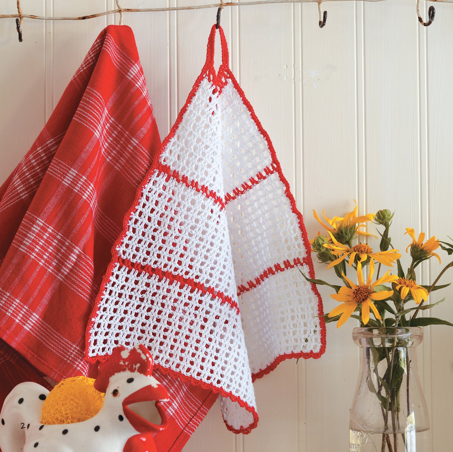 Two towels hang on hooks: one red and white plaid, the other a crocheted white towel with red trim made using the Gooseberry Patch Farmhouse Crochet Digital Download by Leisure Arts. A glass jar with yellow flowers and a ceramic rooster sit nearby against white paneling.