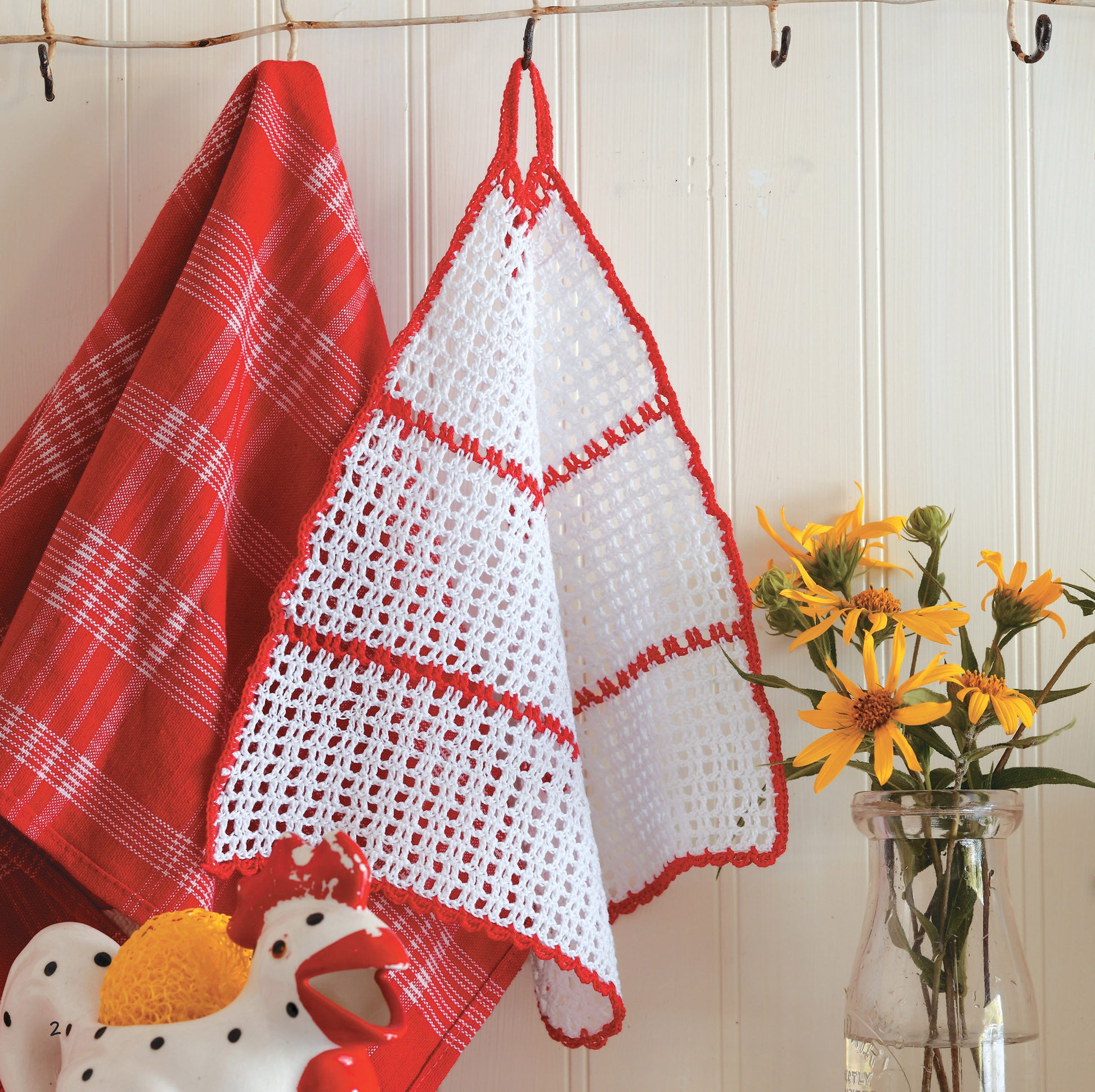 Two towels hang on hooks: one red and white plaid, the other a crocheted white towel with red trim made using the Gooseberry Patch Farmhouse Crochet Digital Download by Leisure Arts. A glass jar with yellow flowers and a ceramic rooster sit nearby against white paneling.