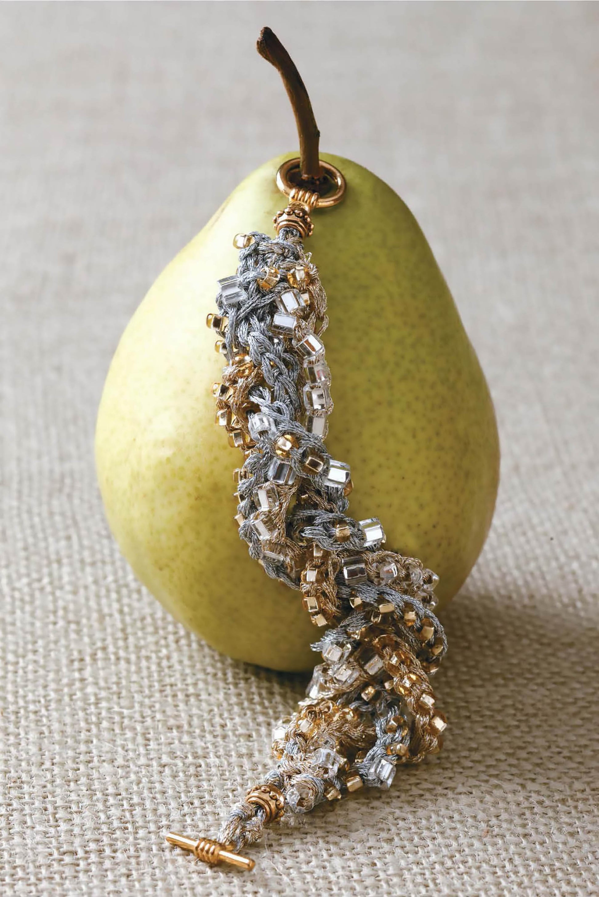 A beaded bracelet from Leisure Arts' Beaded Bracelets to Knit, ideal for beginner jewelry makers, is displayed draped over a green pear resting on a textured beige surface.