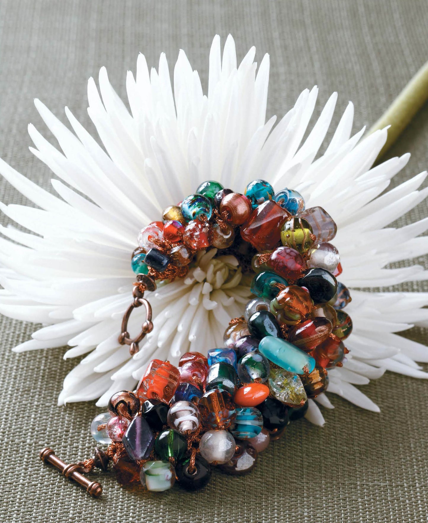 The Beaded Bracelets to Knit kit by Leisure Arts, perfect for beginner jewelry makers, is coiled atop a large white chrysanthemum set against a textured green background.