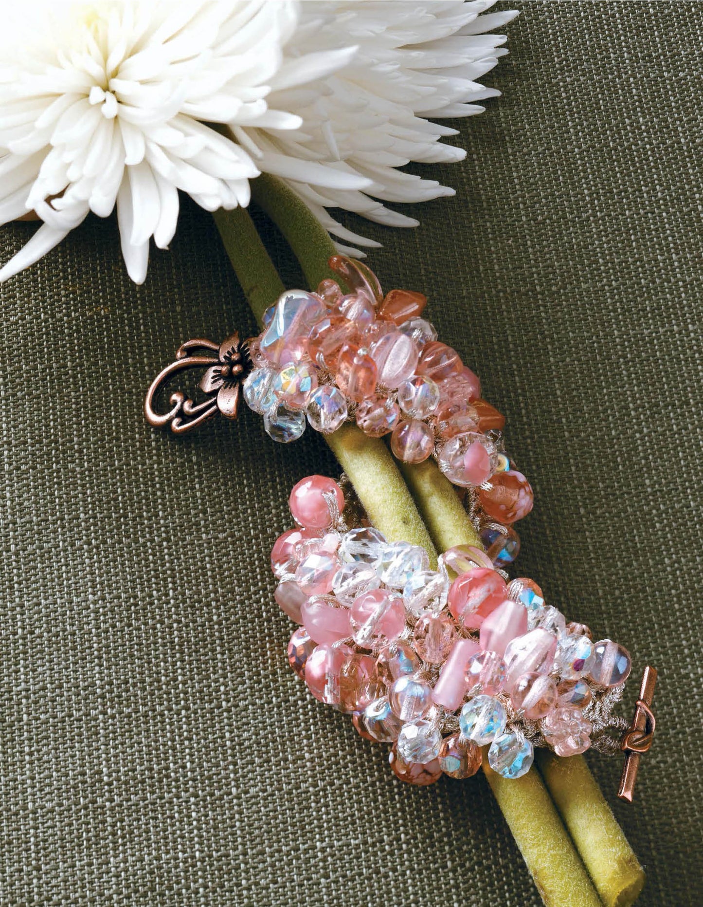 Beaded Bracelets to Knit by Leisure Arts features pink and clear beads with a decorative clasp, displayed on two green stems beside a white chrysanthemum atop textured olive green fabric—perfect for beginner jewelry making.